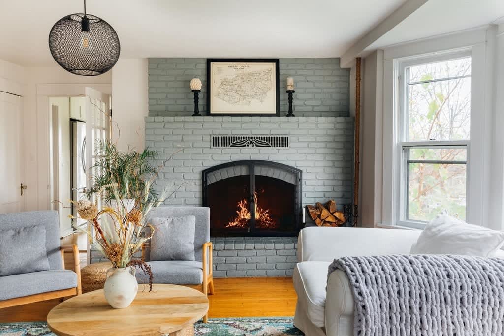 Cozy living room with a light gray painted brick fireplace featuring a black metal insert and a wooden mantel. The room has hardwood floors, a white sofa, gray armchairs, and a round wooden coffee table.