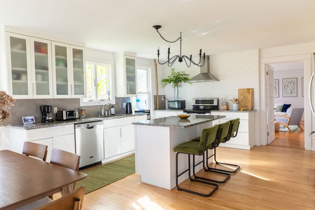 A spacious kitchen features white cabinetry, granite countertops, and stainless steel appliances, including a range hood and dishwasher. A kitchen island with green velvet bar stools serves as a breakfast bar, and a dark chandelier hangs overhead.