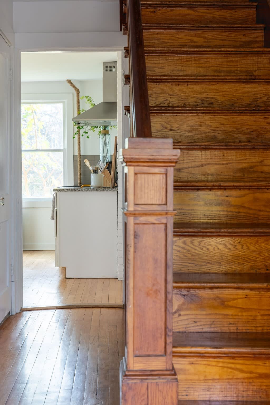 A view of a traditional wooden staircase with a sturdy newel post, looking towards a doorway that opens into the kitchen area. The warm hardwood floors extend throughout the space, suggesting a central entry or hallway.