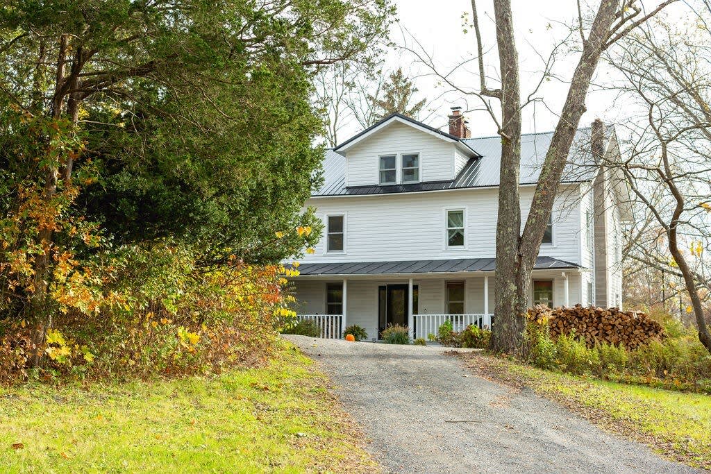 A gravel driveway leads to the front of a white farmhouse, framed by tall trees and green foliage, with a large stack of firewood visible on the right.