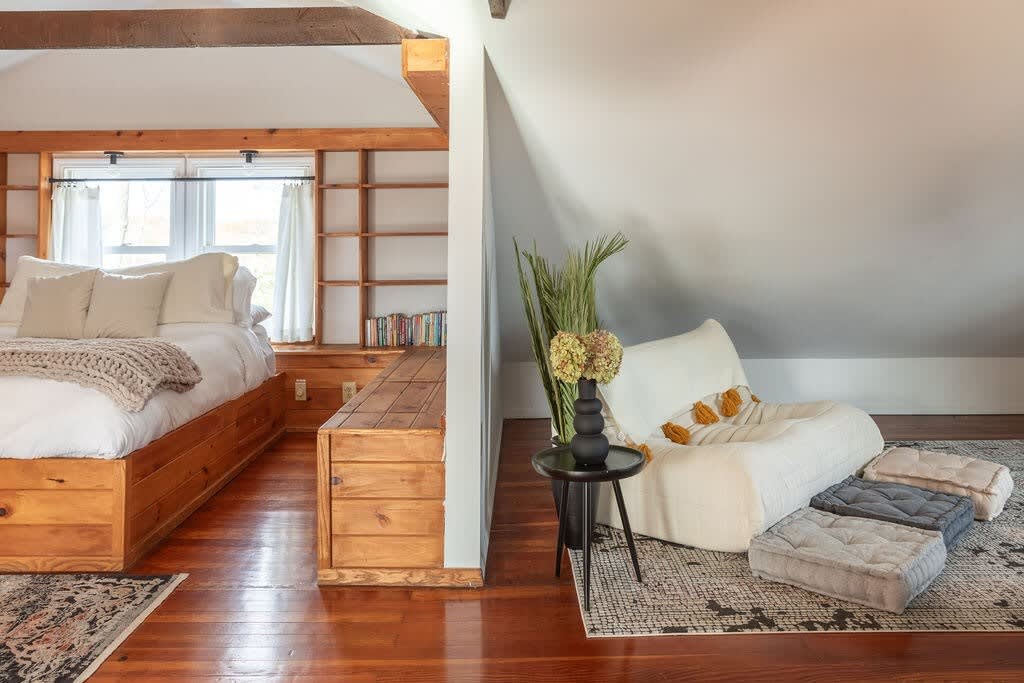 A spacious attic room combines a wooden platform bed with white bedding and a separate lounge area featuring a low white chair and floor cushions on a patterned rug. Exposed wooden beams are visible.