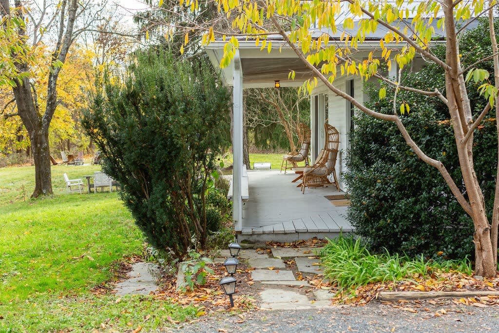 A stone path leads to a white farmhouse porch, partially obscured by lush green bushes and trees with yellow autumn leaves, revealing a glimpse of outdoor seating.