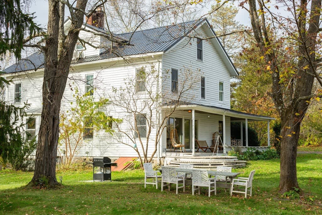 The back of a white farmhouse features a covered porch with seating, a BBQ grill, and an outdoor dining set on a green lawn, surrounded by mature trees.