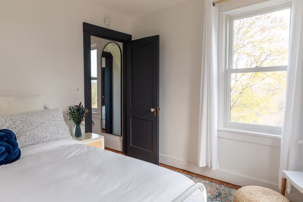 A bright bedroom features a white bed with a blue decorative pillow, light wood floors, and a dark arched mirror reflecting a dark door. White curtains frame a large window with a view of trees.