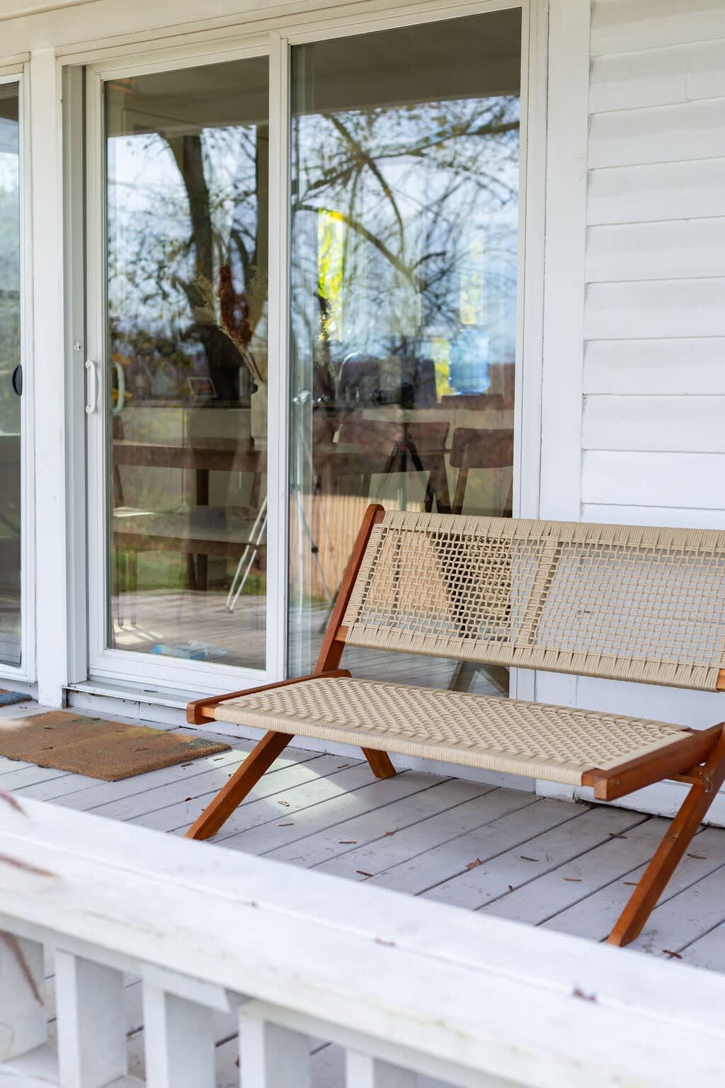A modern wooden bench with woven seating sits on a white wooden porch, next to large sliding glass doors reflecting the outdoor scenery.