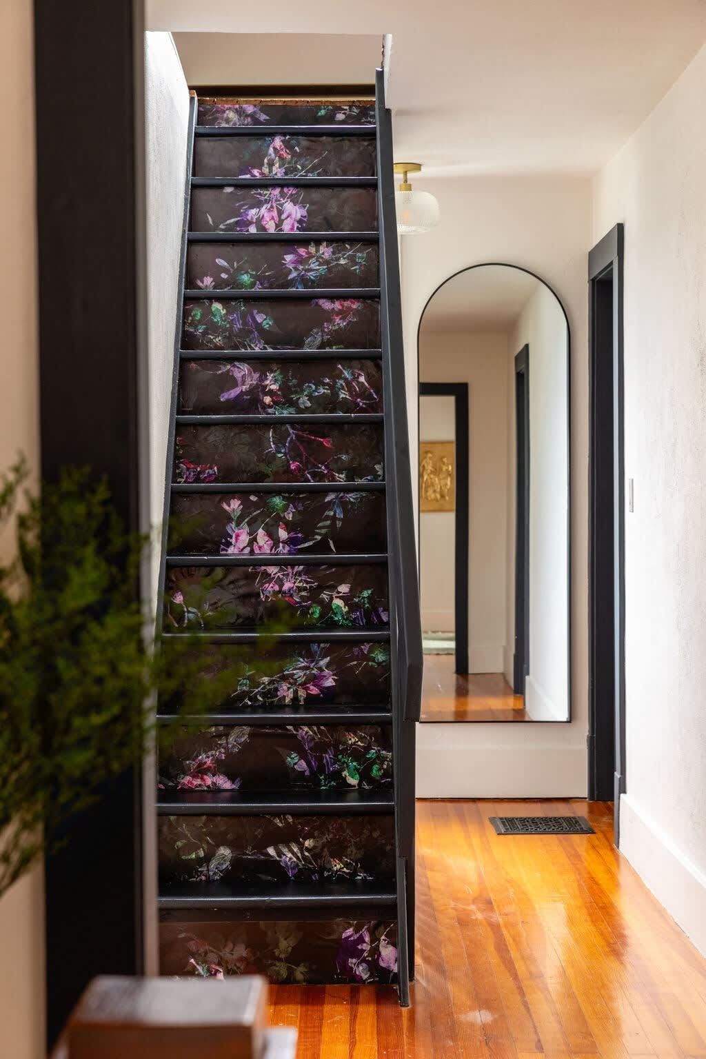 A striking staircase with dark risers adorned with vibrant floral wallpaper leads upstairs, complemented by polished hardwood floors and a tall arched mirror in the hallway.