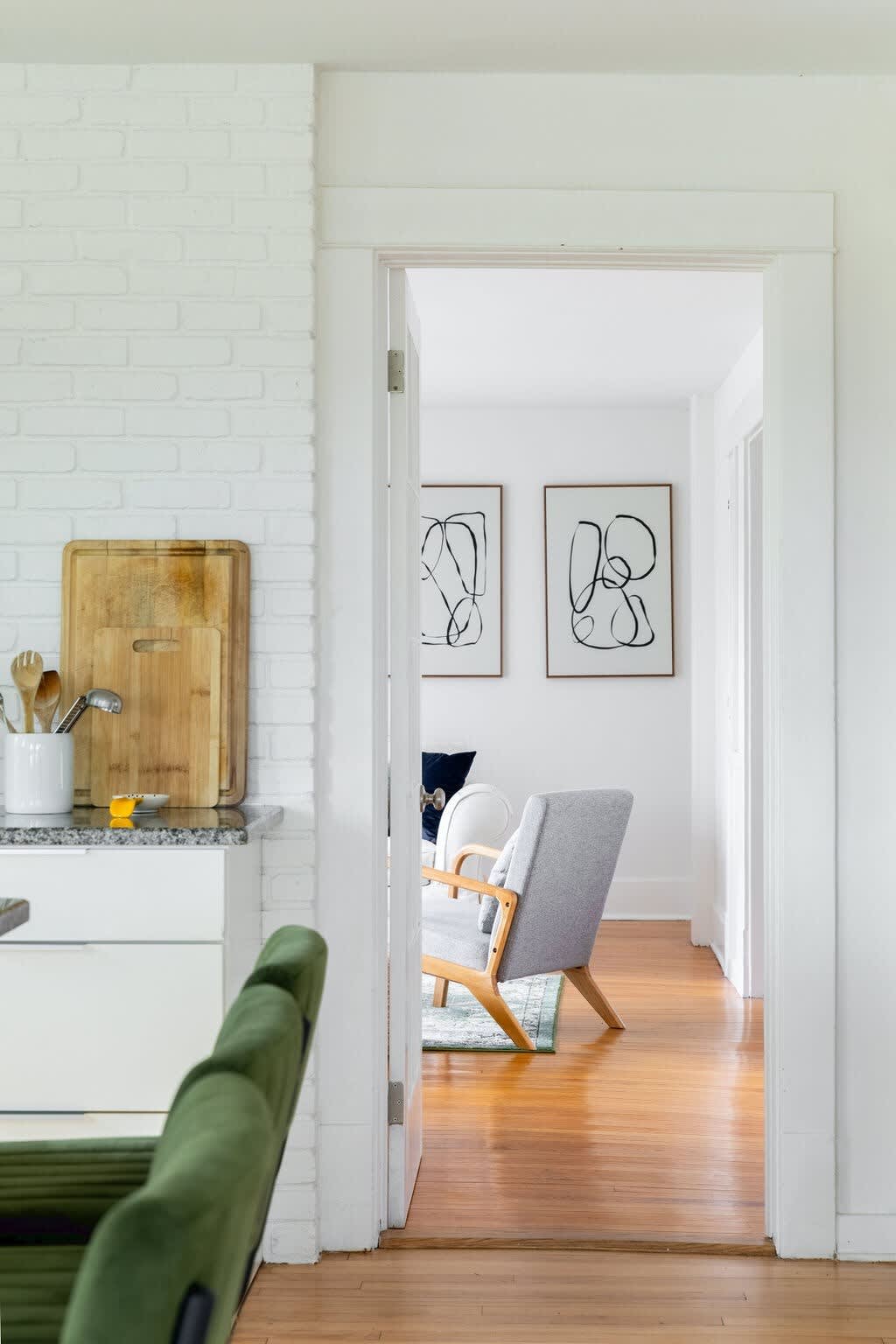 A doorway frames a view into the living room, revealing a gray armchair, a white sofa, and two framed abstract artworks on a white wall. The room features warm hardwood floors and a green patterned rug.