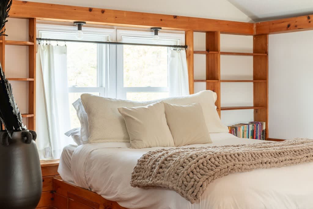 A close-up of the wooden platform bed, adorned with white bedding and a chunky knit throw, is framed by built-in wooden shelving around a window. The room features exposed wooden beams.