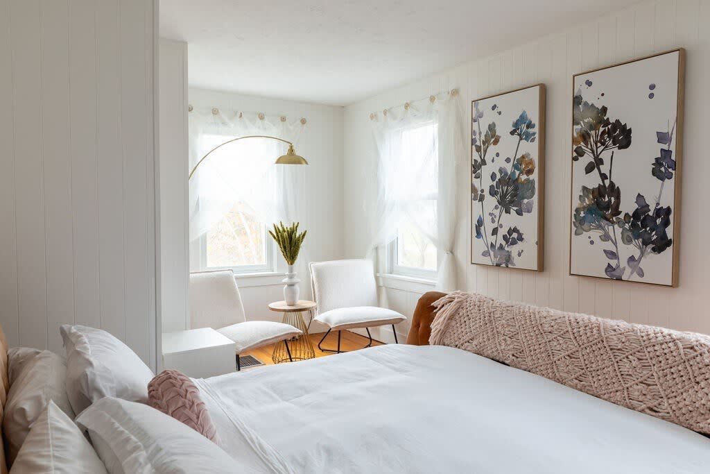 Bright bedroom with white paneled walls, a large bed with white bedding, and a cozy corner seating area with two white chairs, a small table, and a brass floor lamp. Two large floral artworks adorn the wall.