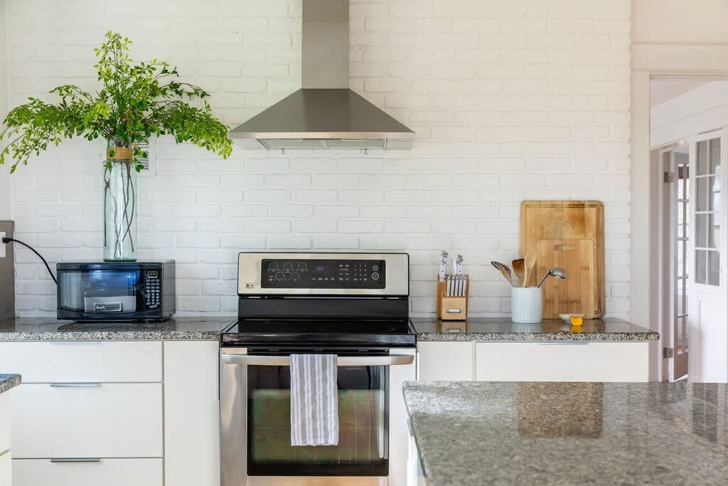 A section of the kitchen features a stainless steel range and matching hood against a white painted brick wall. Granite countertops flank the stove, holding a microwave, cutting boards, and kitchen utensils.