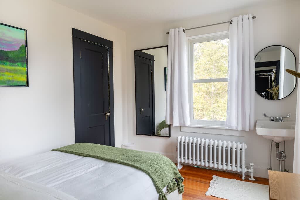 A bright bedroom with white walls, a bed with white bedding and a green throw blanket, featuring a large window with white curtains and a white radiator below. A full-length mirror and a wall-mounted sink are also visible.