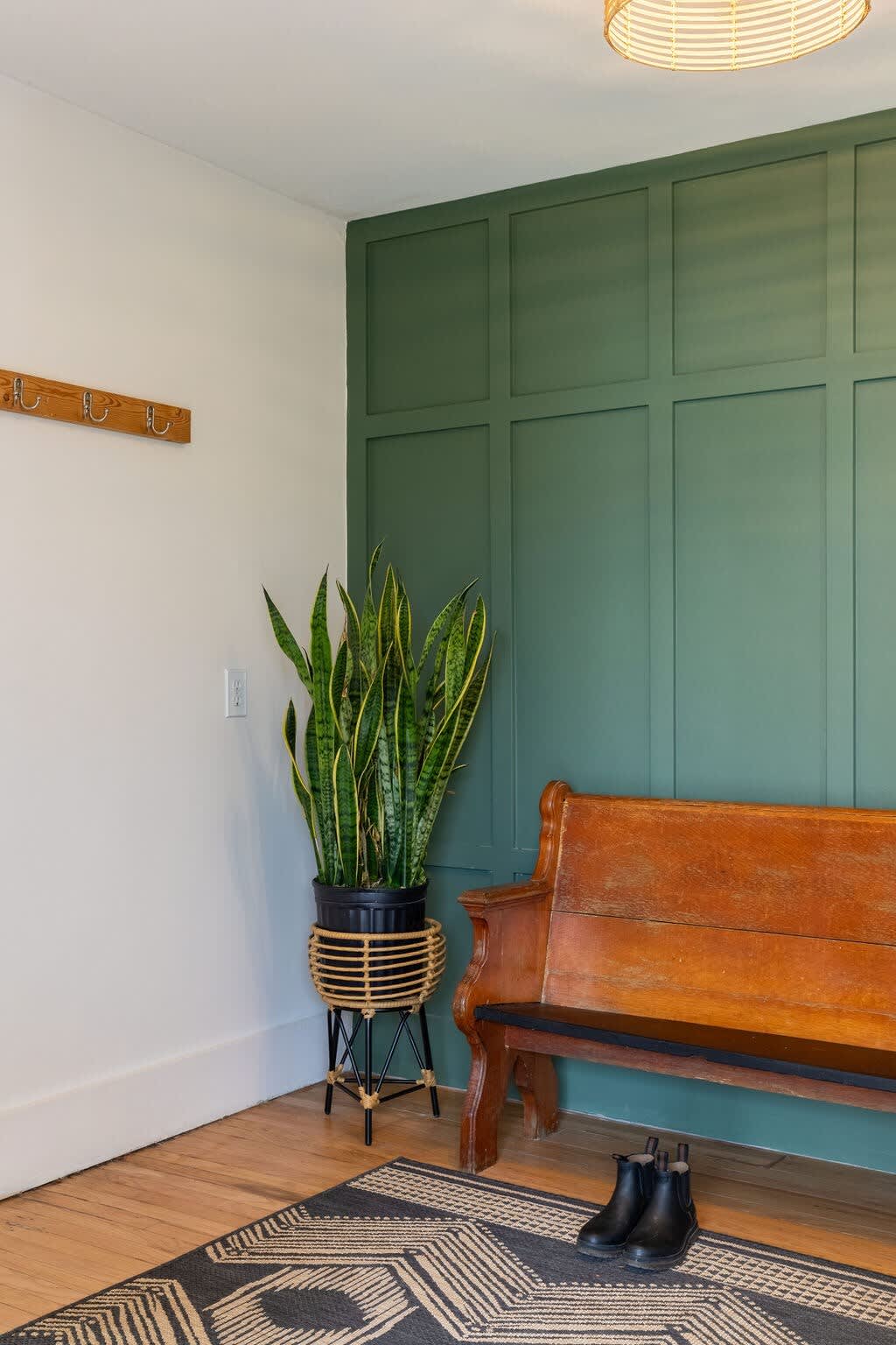 A welcoming entryway features a dark green paneled accent wall with a wooden bench and a snake plant in a woven planter. A wooden coat rack is on the adjacent white wall, and a geometric patterned rug covers the hardwood floor.