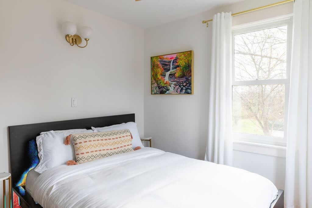 A serene bedroom with white walls, a dark headboard, white bedding, and a decorative lumbar pillow, illuminated by natural light from a large window with white curtains and a gold curtain rod. A colorful waterfall painting hangs above the bed.