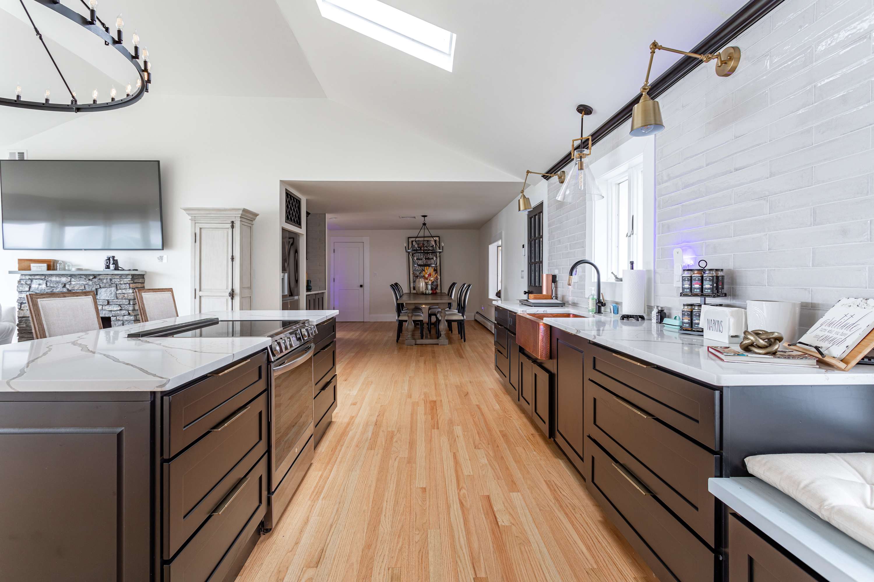 Detailed view of the modern kitchen, showing dark cabinetry, white countertops, a copper sink, and a large island with a built-in range, extending towards a dining area in the background.