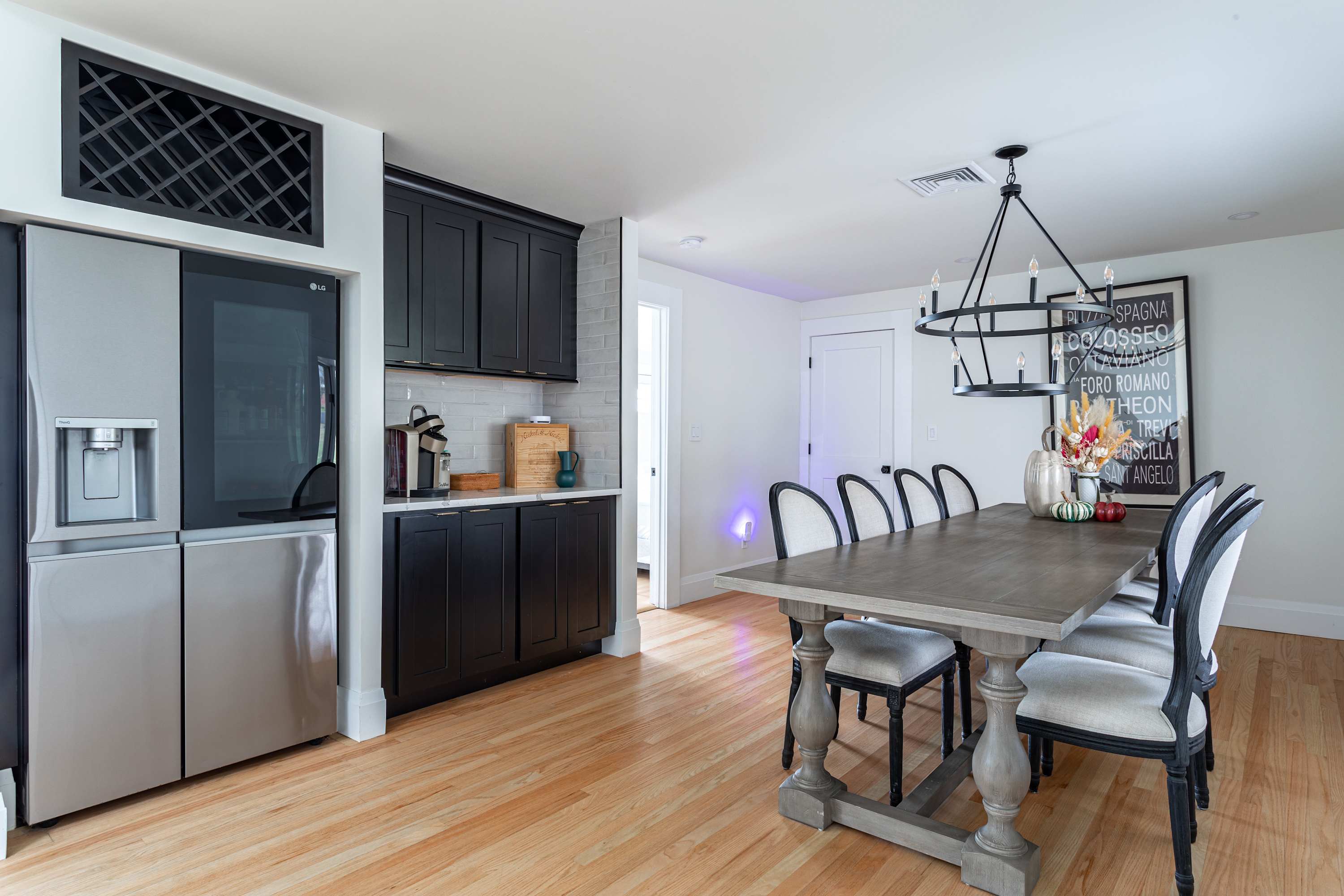 Elegant dining area featuring a rustic wooden table with upholstered chairs, a large black chandelier, and a built-in cabinet with a wine rack, adjacent to a modern stainless steel refrigerator.