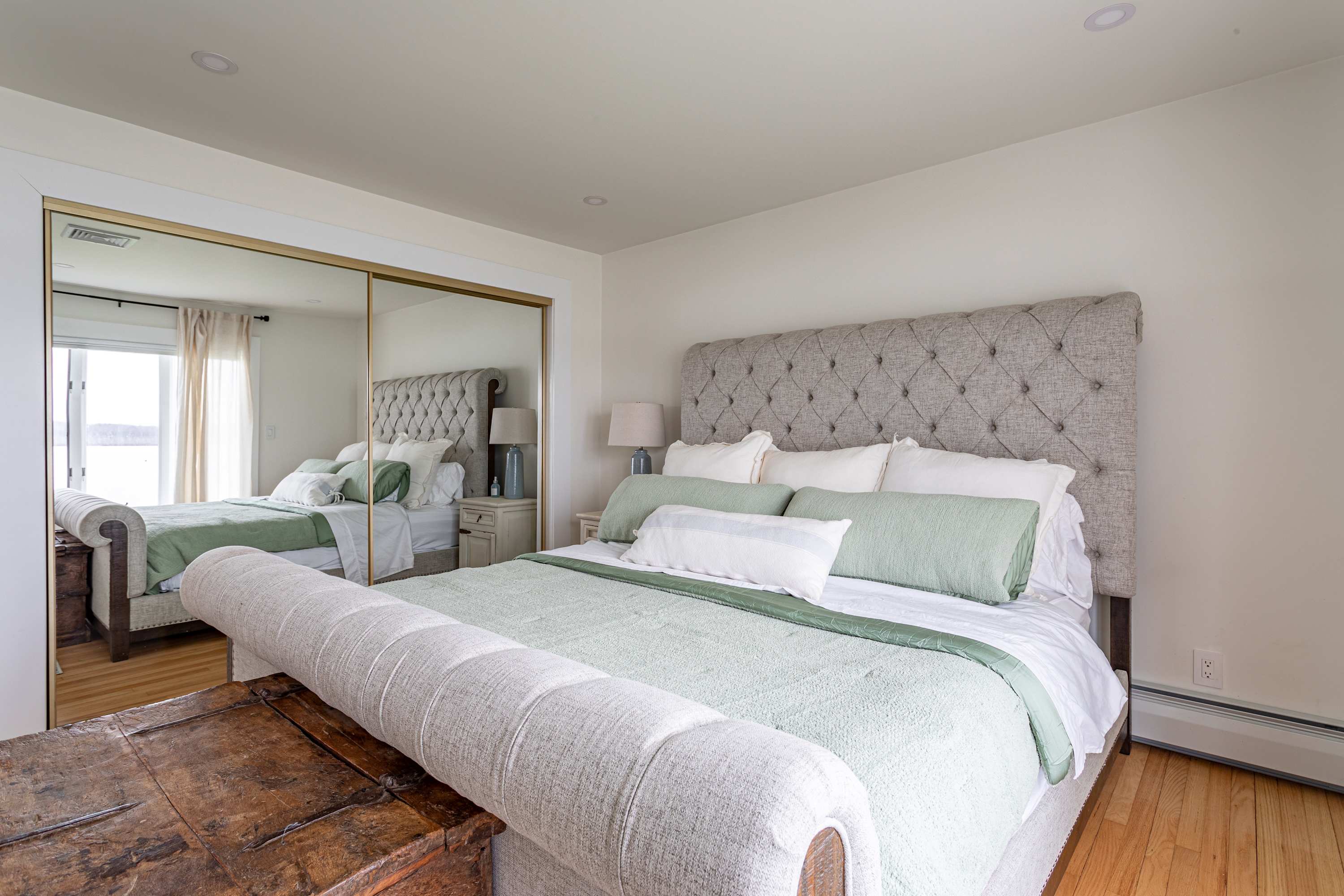 A bright bedroom featuring a large bed with a light gray tufted headboard, light green and white bedding, and a wooden chest at the foot of the bed. A mirrored closet reflects the room and a window with a water view.