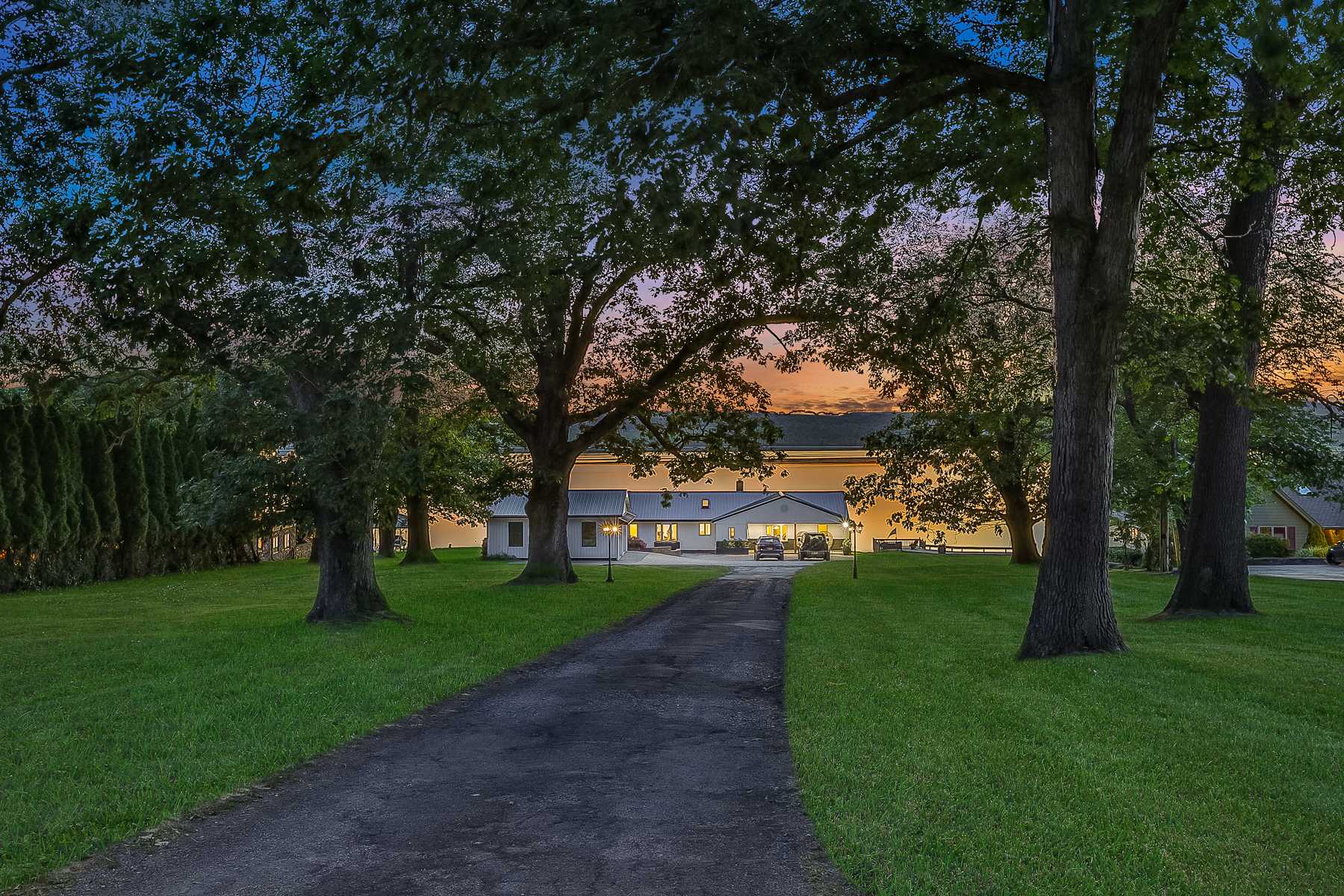 Long asphalt driveway leading to the lakehouse, framed by mature trees and manicured lawns, with the house visible in the distance under a twilight sky.