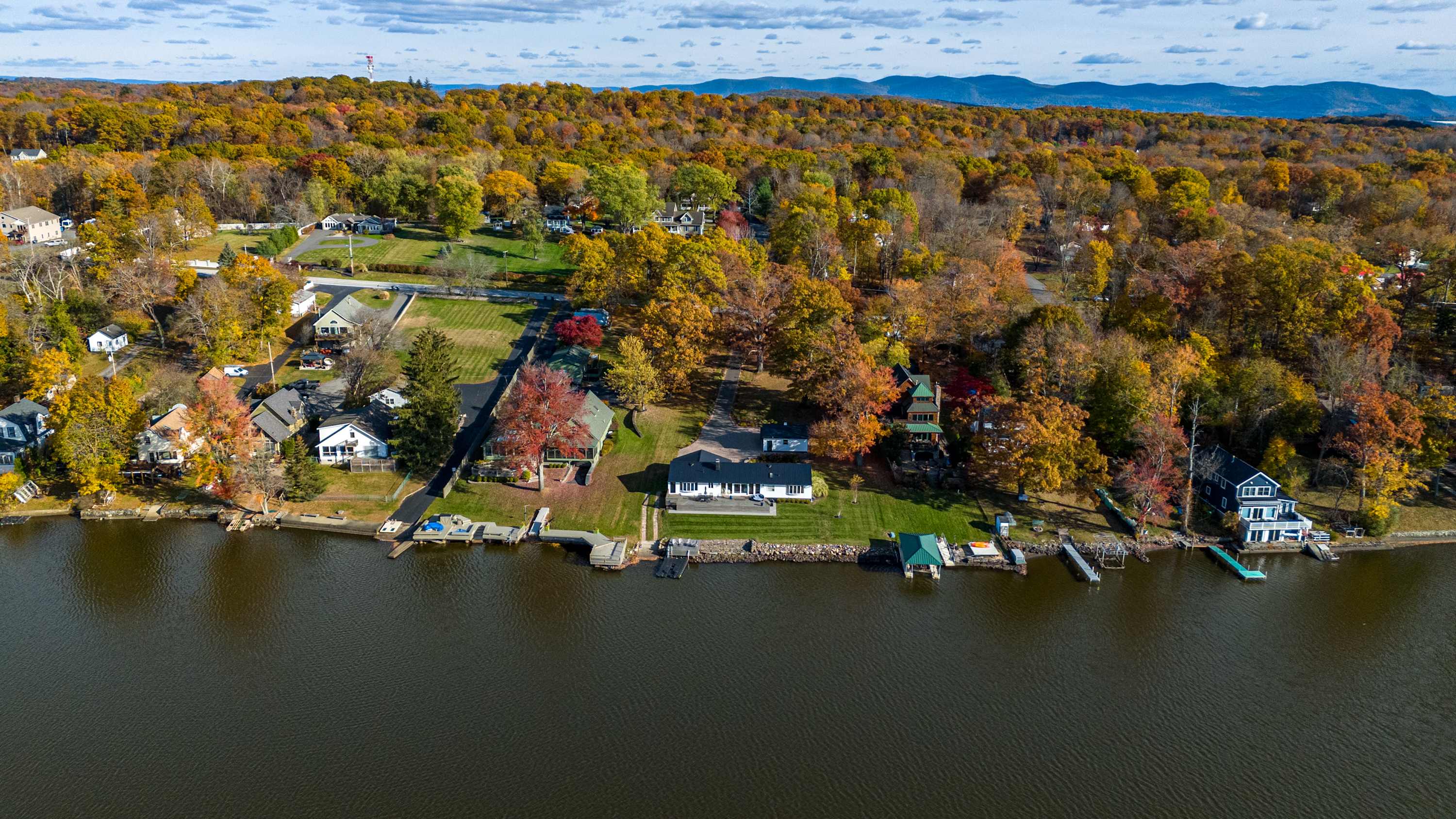 An aerial view showcasing a lakefront property with a white house and green lawn, surrounded by vibrant autumn trees. Several docks extend into the calm lake, with other houses visible along the shore.