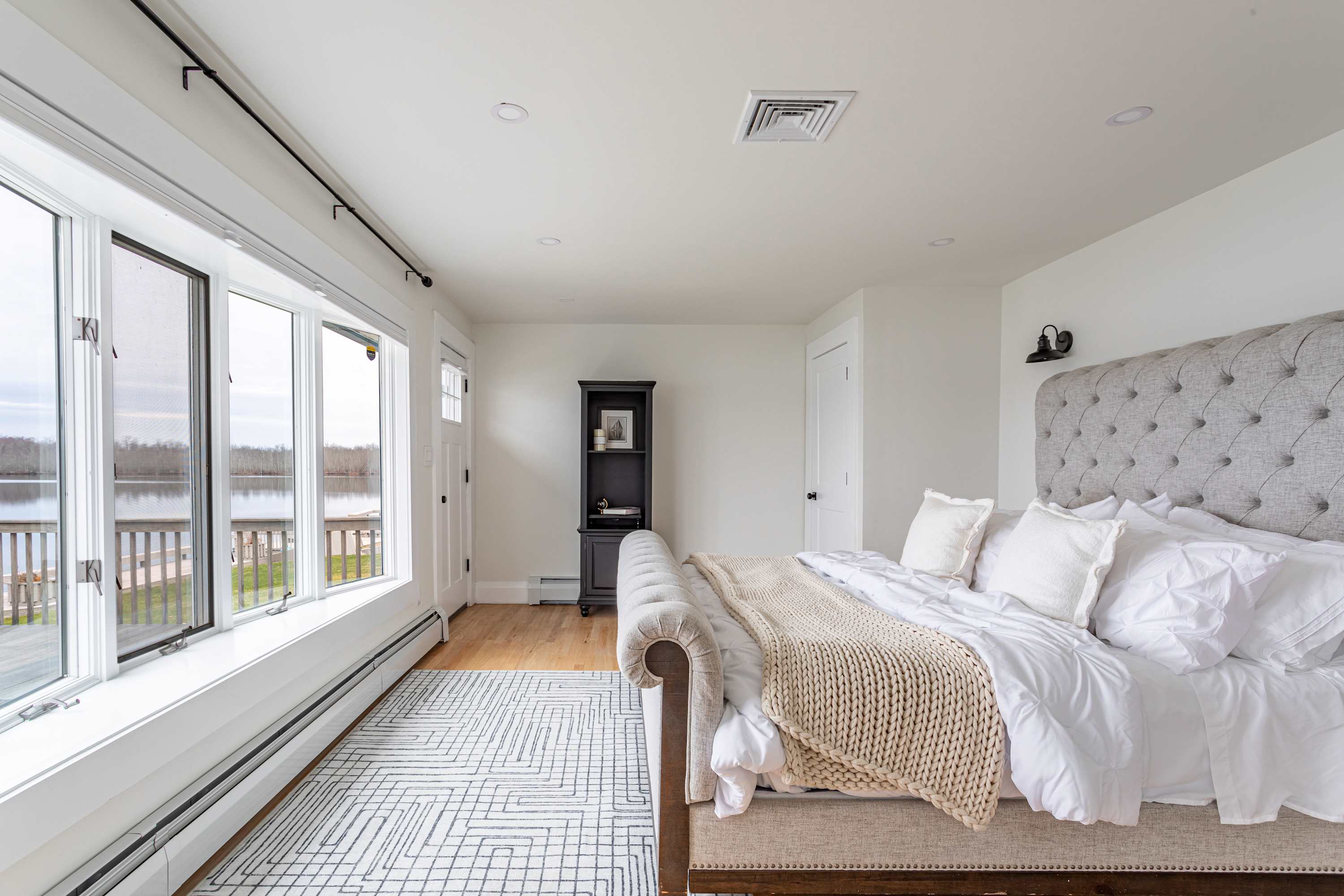 Another view of the bedroom, showing the tufted upholstered bed, white bedding, and a large window with lake views, along with a black bookshelf and a door leading to the bathroom.