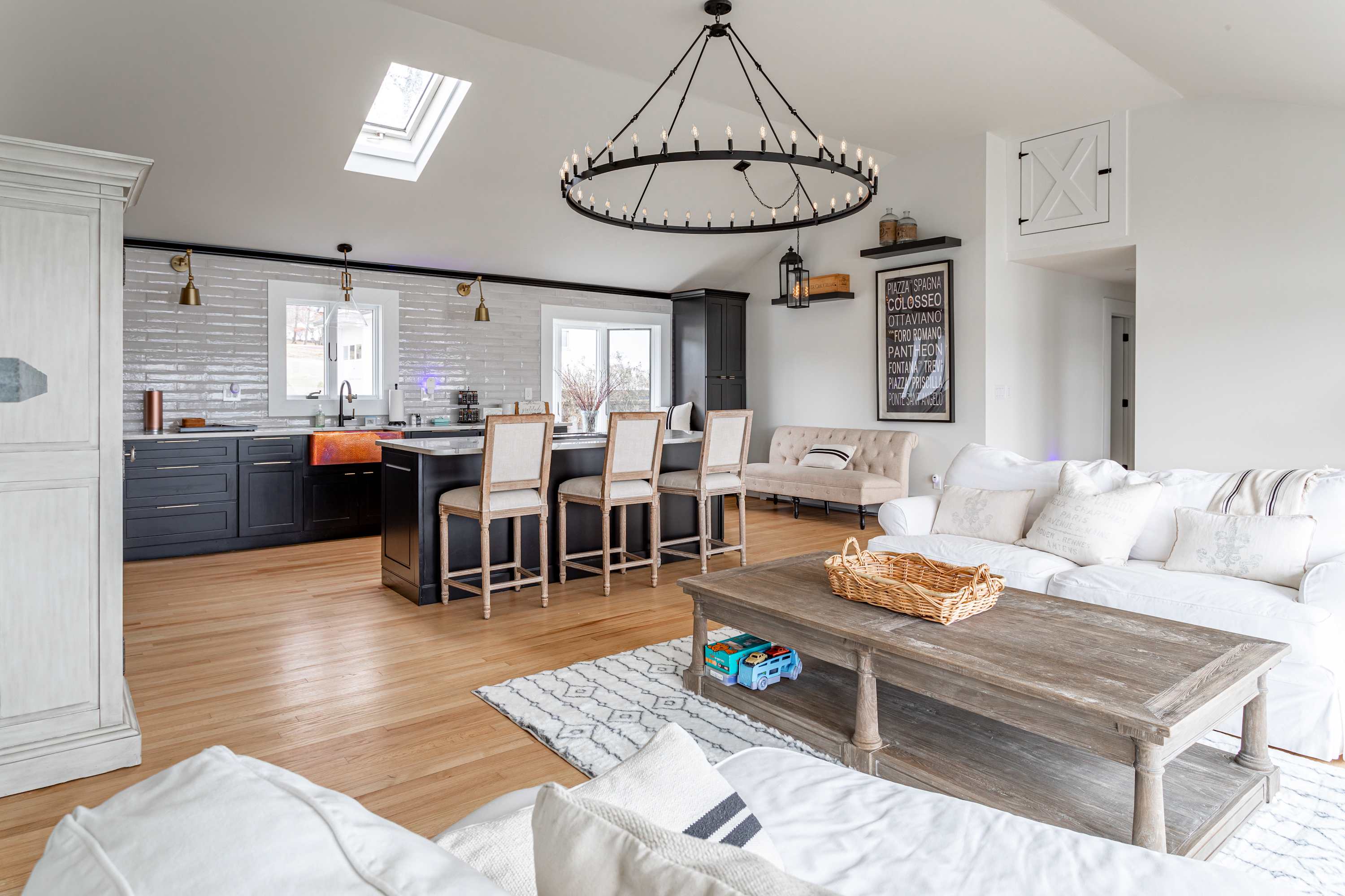 Open-concept living space featuring a modern kitchen with dark cabinetry, a large island with seating, and a living area with a white sofa and wooden coffee table, all under a vaulted ceiling with a large circular chandelier.