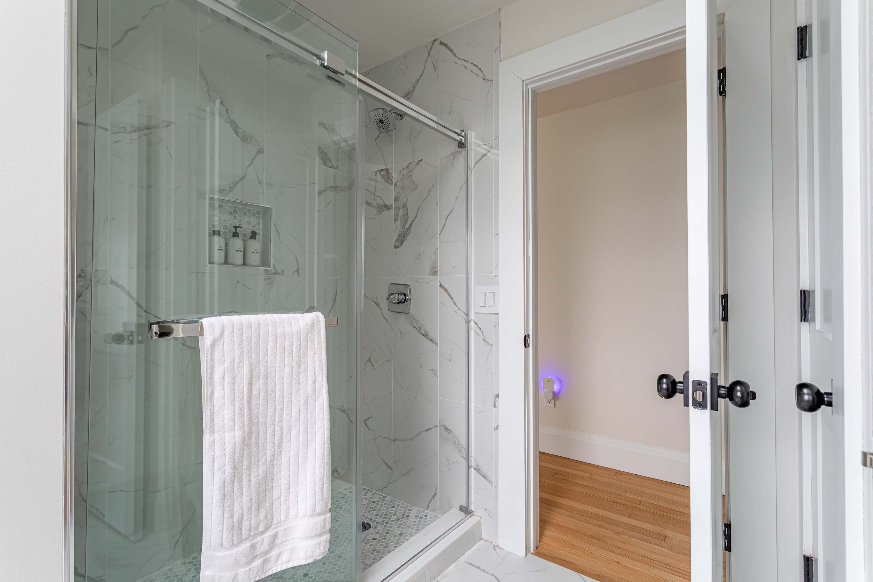 A modern bathroom featuring a spacious glass-enclosed shower with white marble-patterned tile walls and a mosaic tile floor. A white towel hangs on the shower door handle.