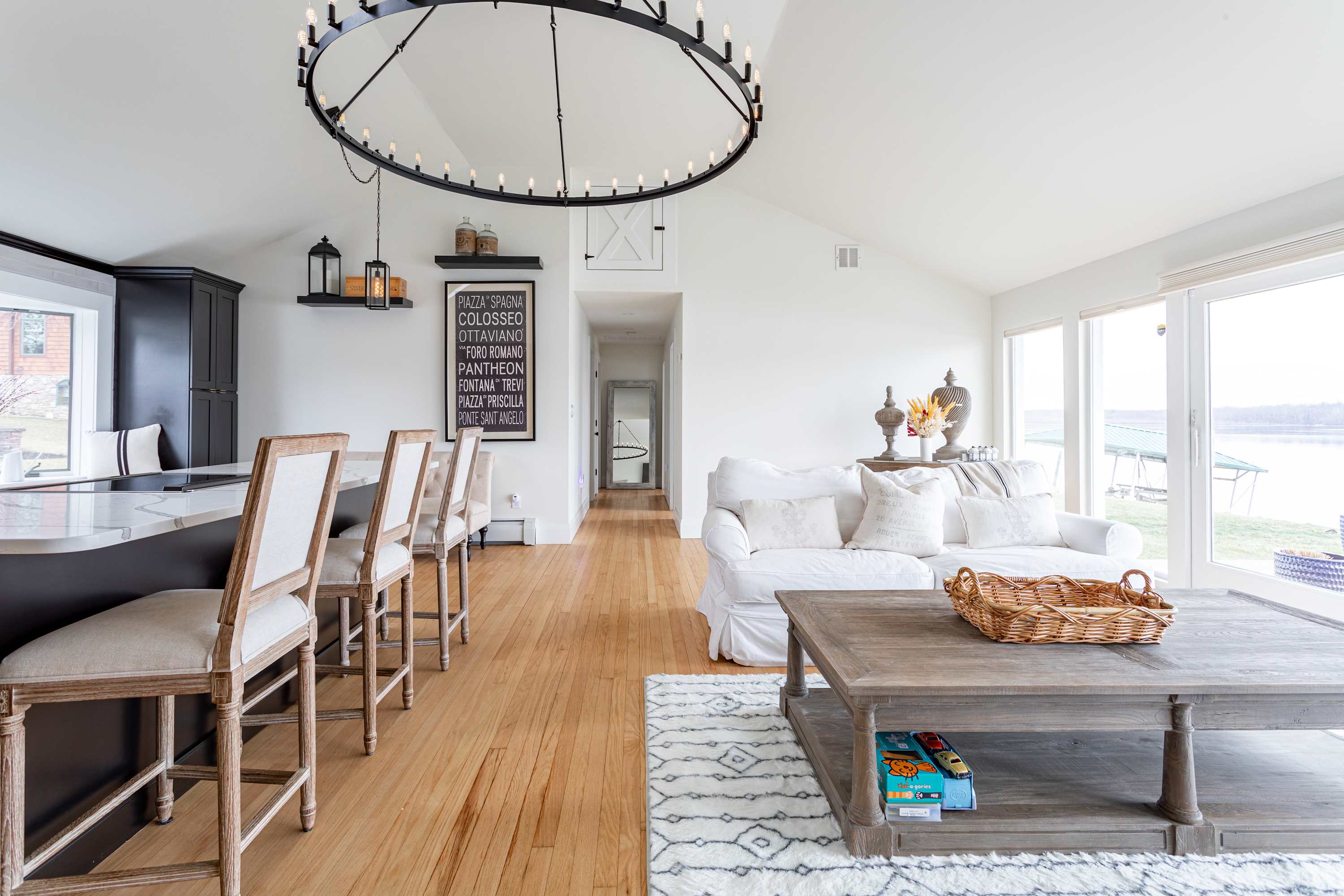 Open-concept living space featuring a white slipcovered sofa, a large circular chandelier, and a breakfast bar with dark cabinetry and bar stools, all with hardwood floors and lake views.