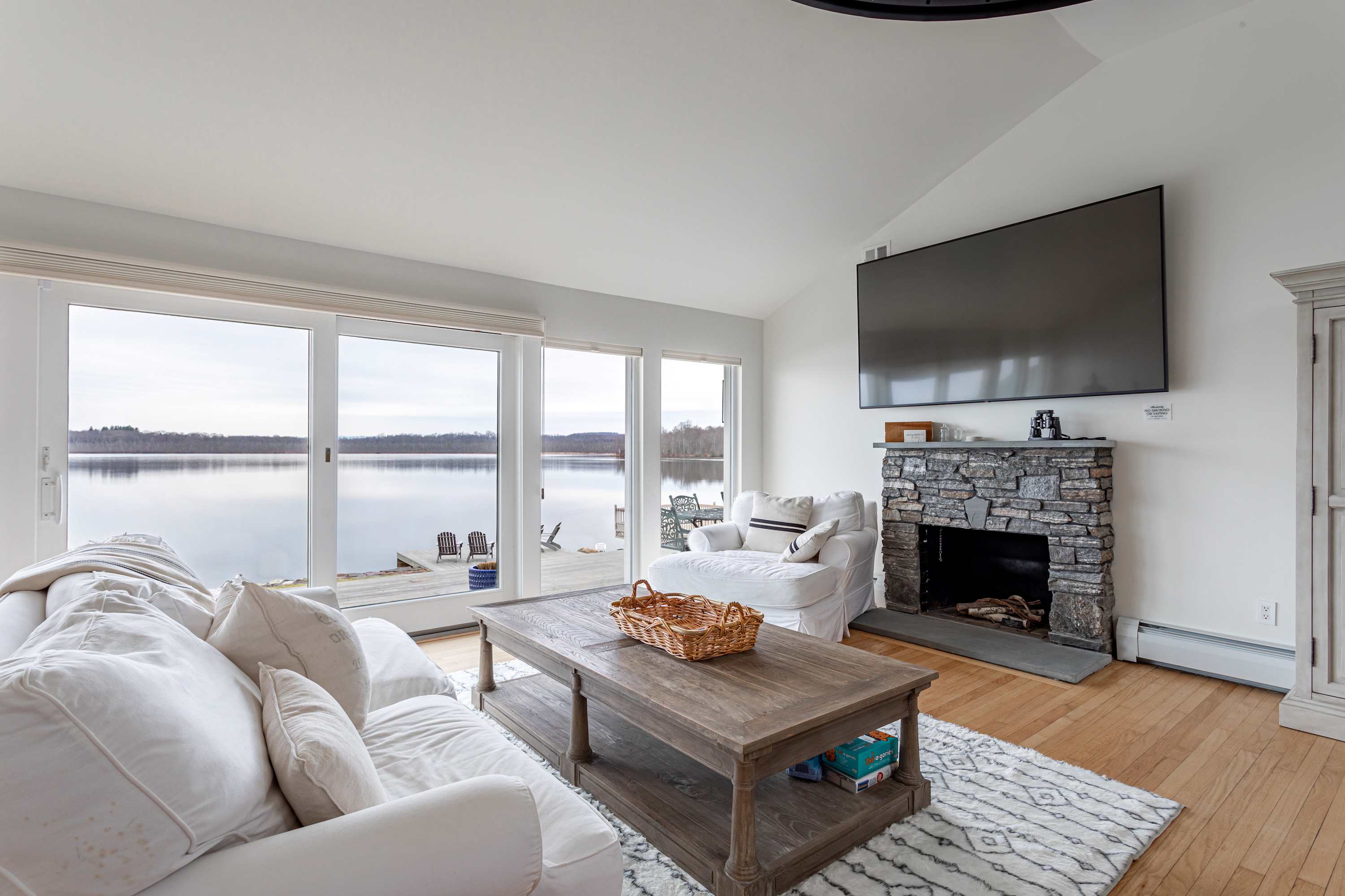 Cozy living room with a white slipcovered sofa, a rustic wooden coffee table, a stone fireplace with a mounted TV, and large sliding glass doors opening to a deck with lake views.