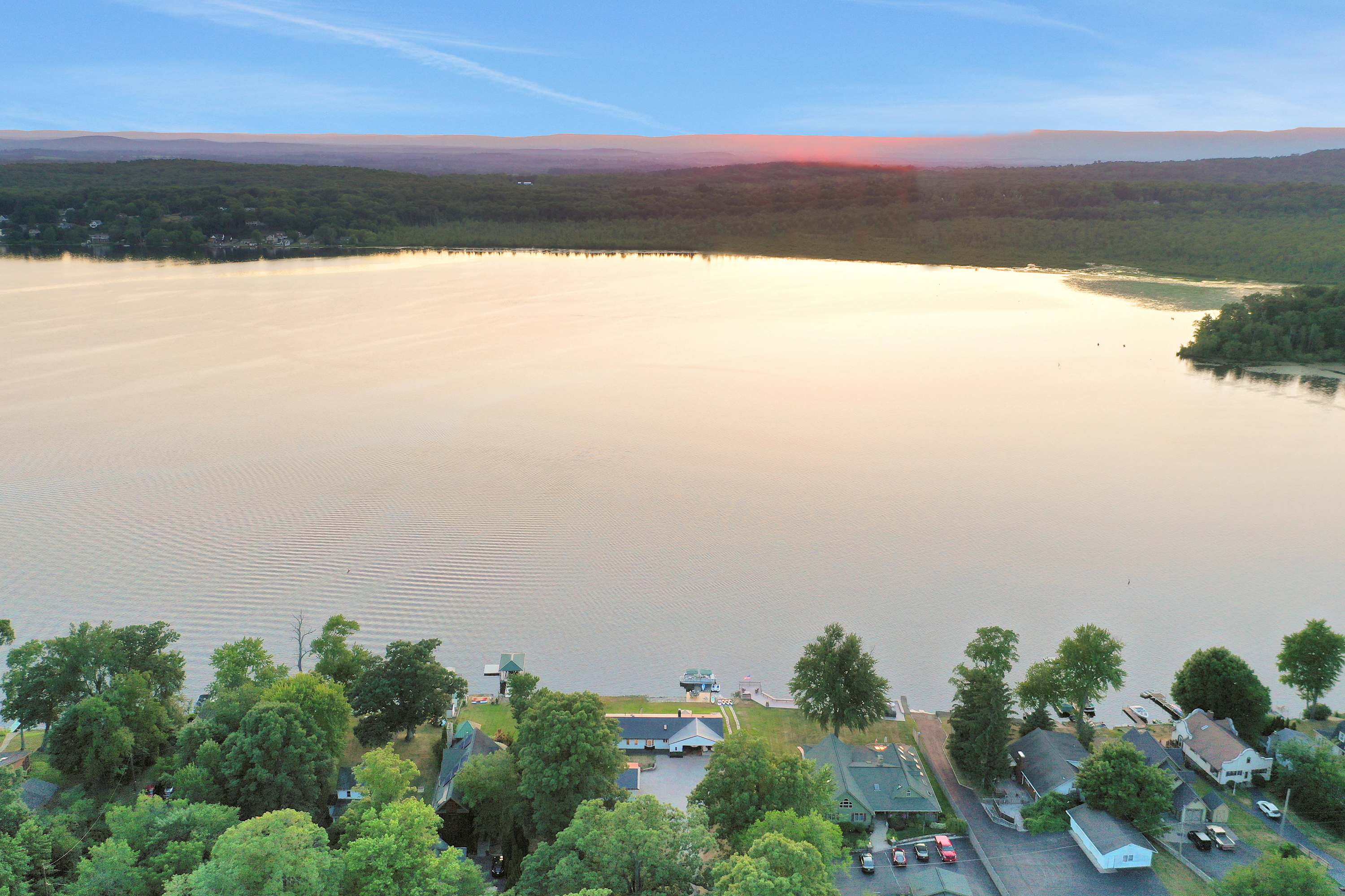 Expansive aerial view of a large, calm lake surrounded by lush green trees and residential properties along the shoreline, under a soft, golden sunset sky.