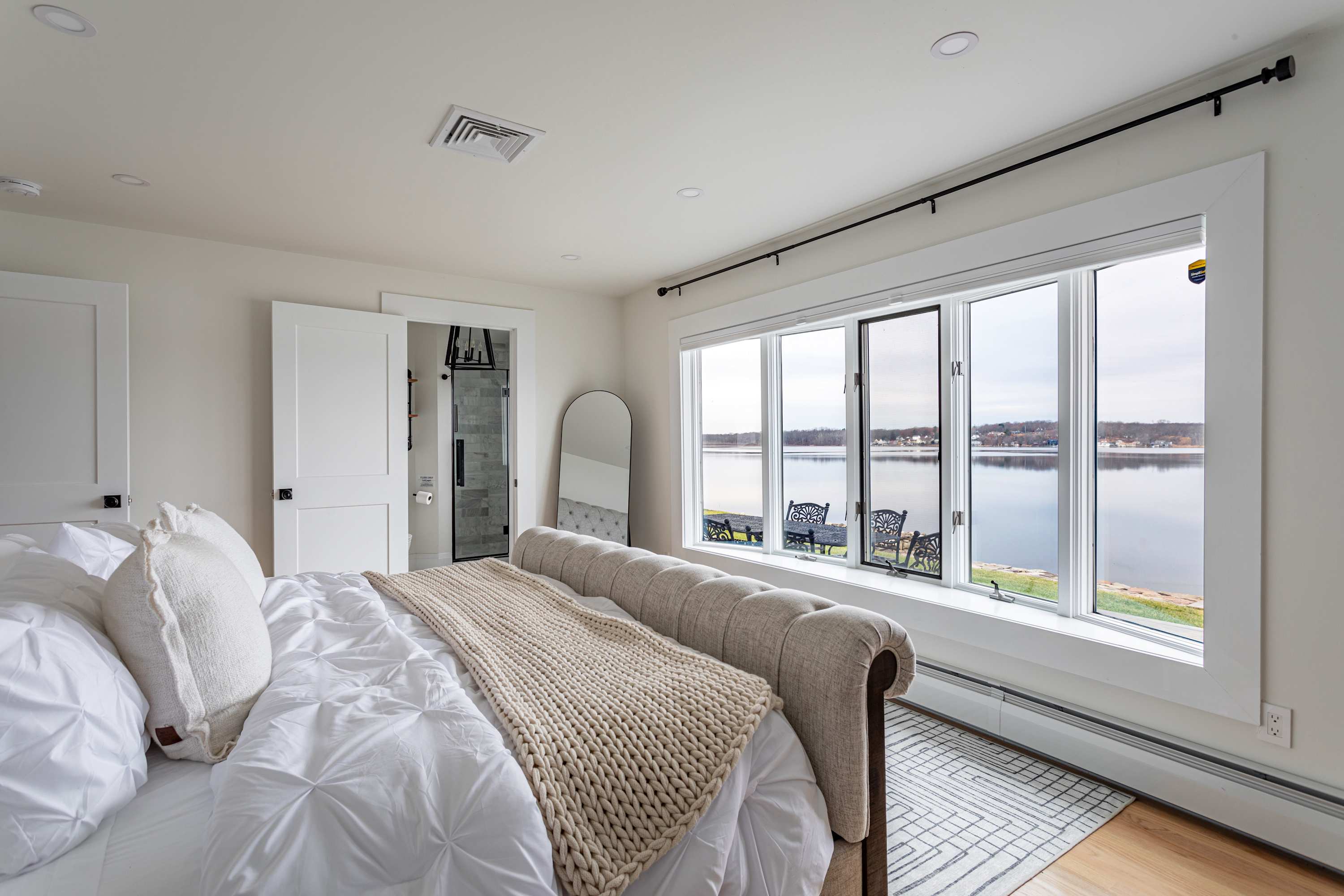 Bright bedroom with a tufted upholstered bed, white bedding, and a large window offering expansive lake views, featuring hardwood floors and a simple, clean aesthetic.