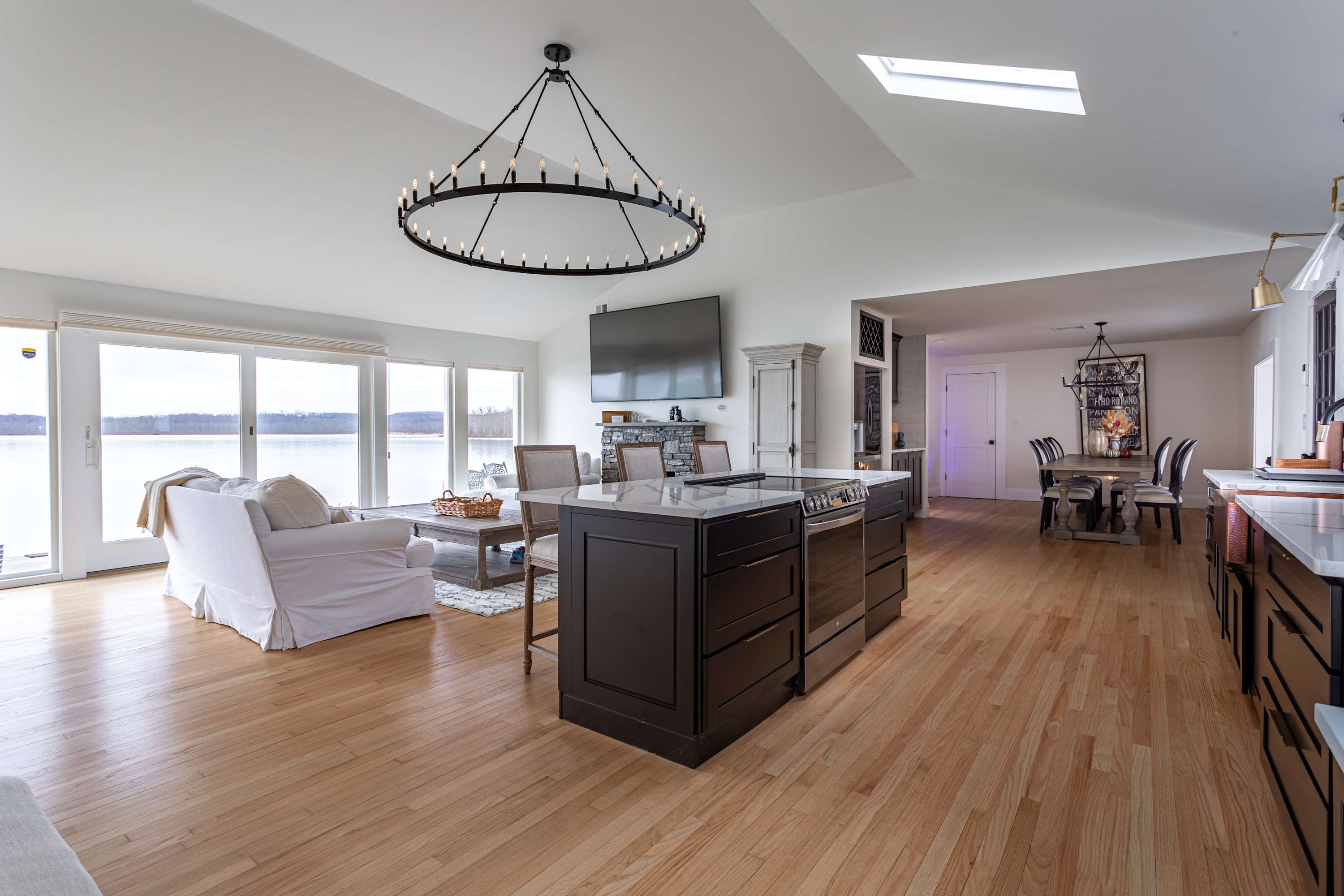 Expansive view of the open-concept living space, kitchen, and dining area, featuring a white sofa, stone fireplace, kitchen island, and dining table, all with panoramic lake views through a wall of windows.
