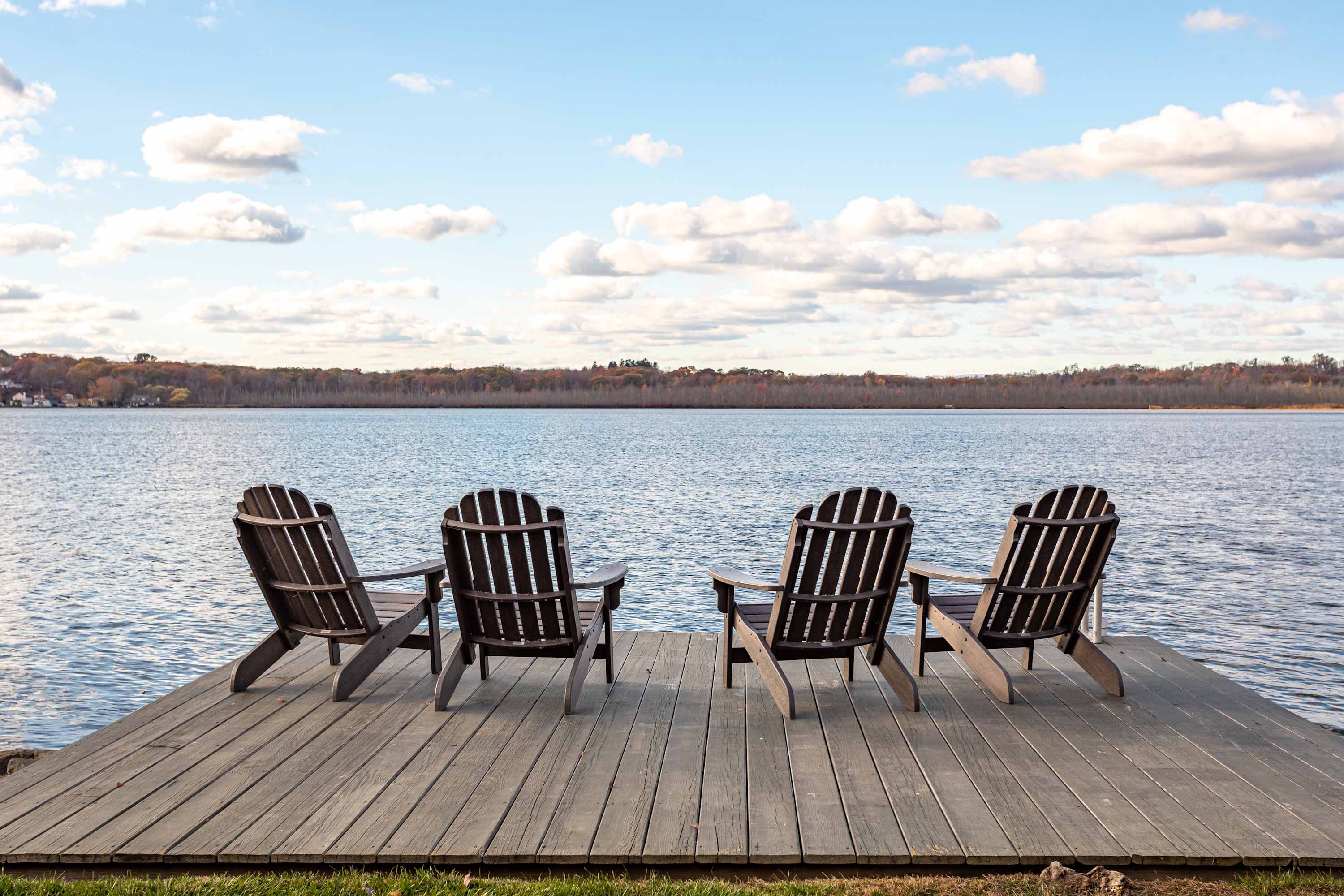 Wooden dock extending into a calm lake, featuring four dark Adirondack chairs arranged for enjoying the serene water views under a partly cloudy sky.