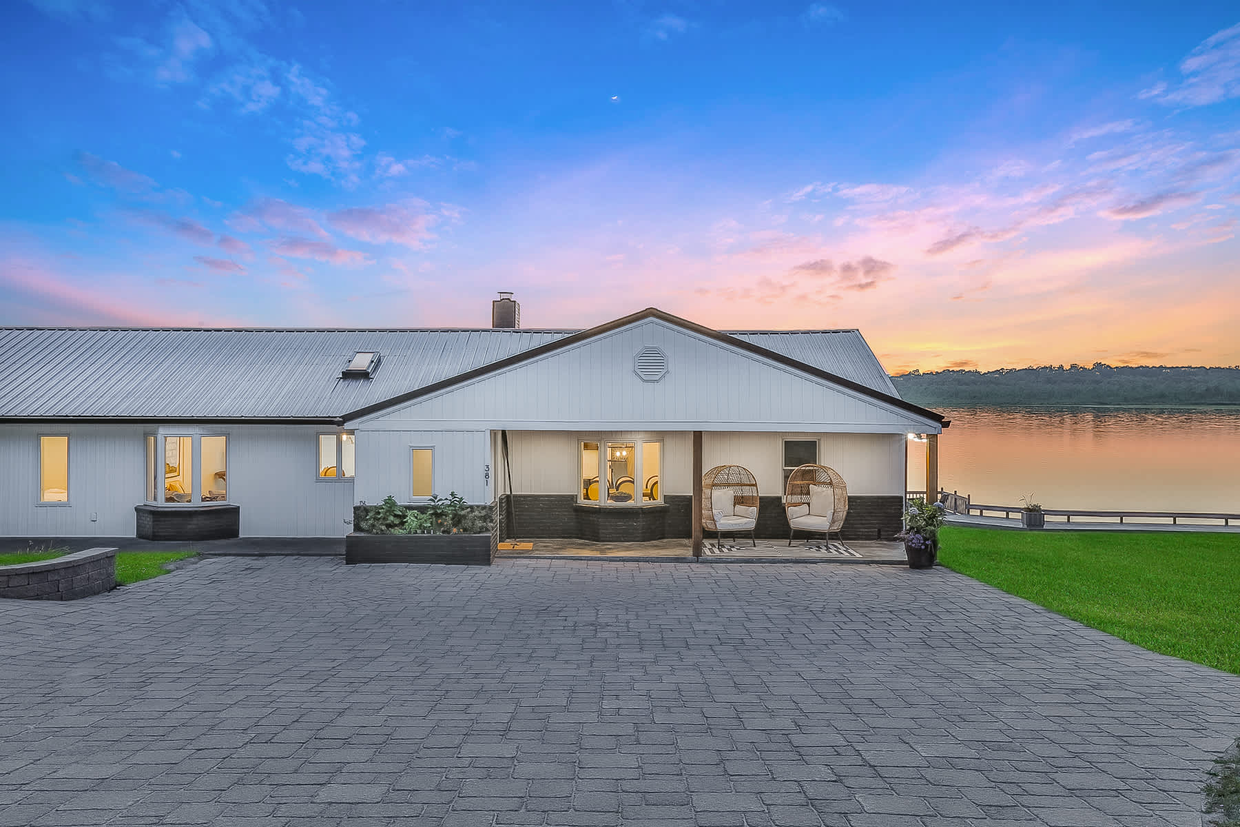 The front exterior of a white lakehouse at dusk, featuring a paved driveway, a covered porch with two hanging egg chairs, and a view of the lake under a colorful sunset sky.