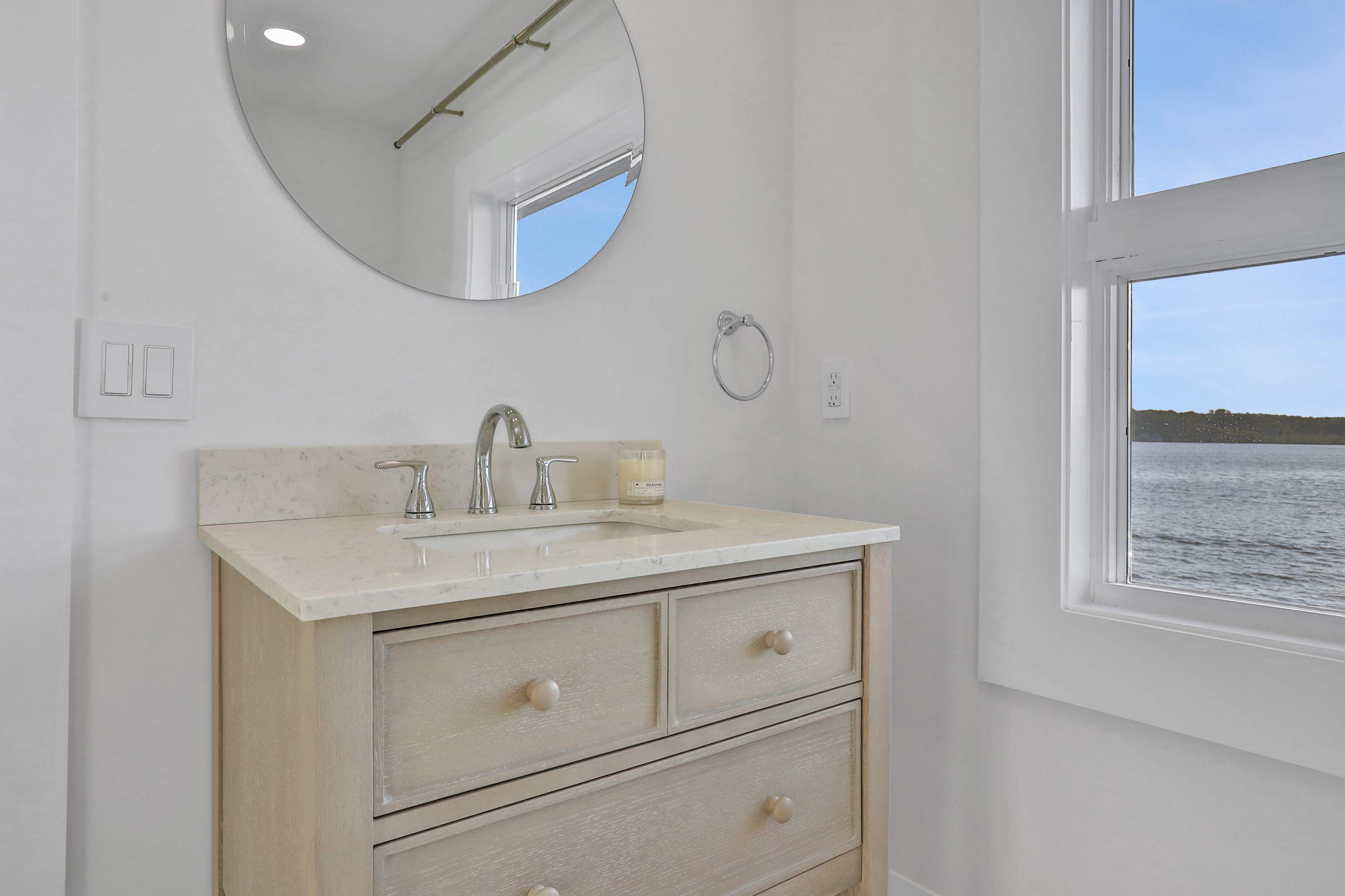 A close-up of a light wood vanity with a white marble countertop and a modern chrome faucet. A round mirror hangs above, and a window provides a clear view of the lake.