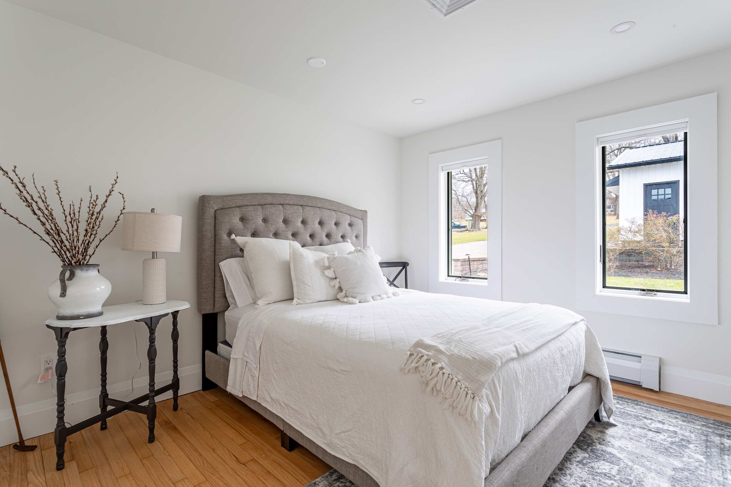 A bright bedroom featuring a gray tufted queen bed with white bedding, light hardwood floors, and two windows. A small round table with a lamp and vase of branches serves as a nightstand.