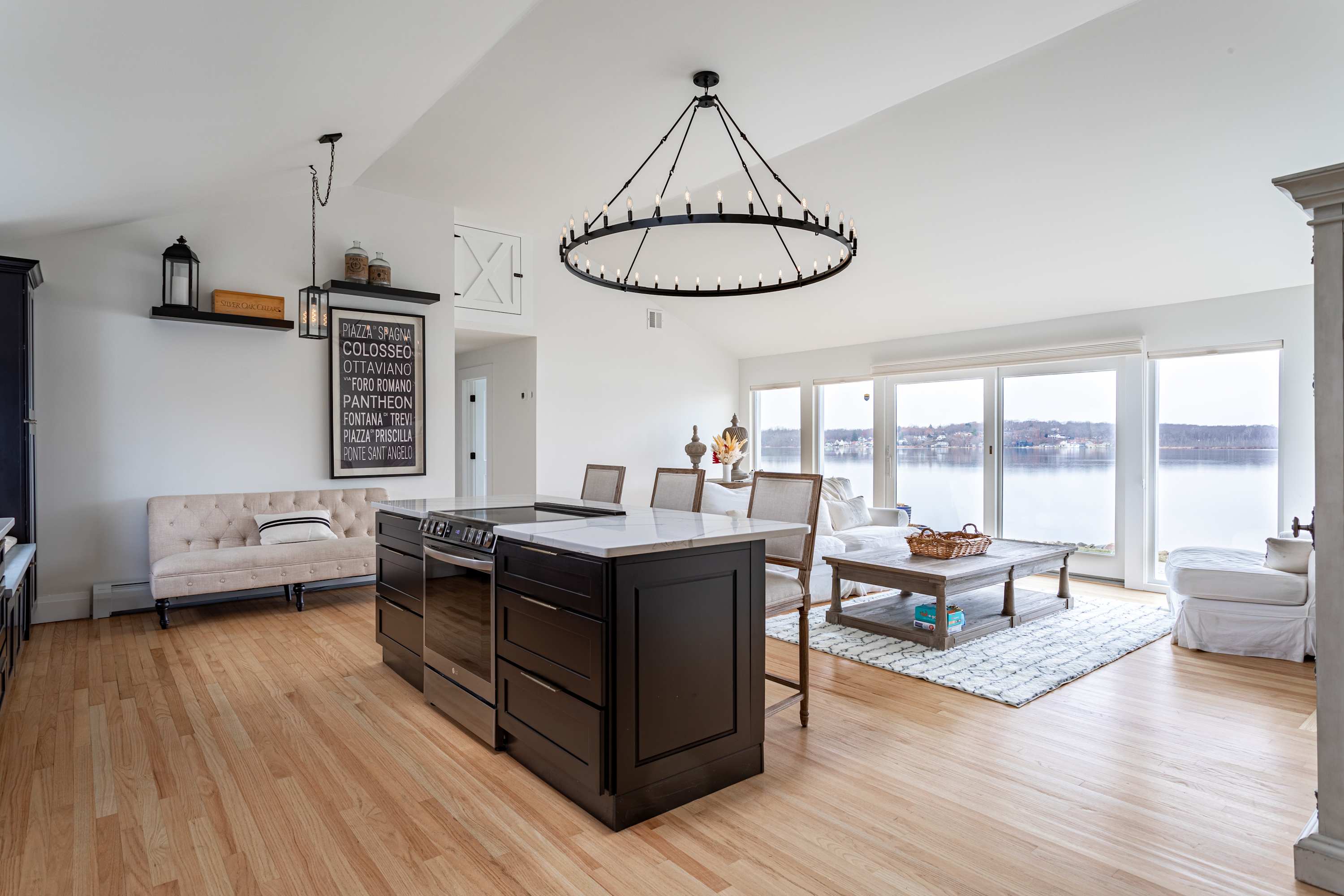 View of the open-concept living and kitchen area from a different angle, highlighting the kitchen island with seating, a small beige sofa, and the living room with a white sofa and coffee table, all with lake views through large windows.