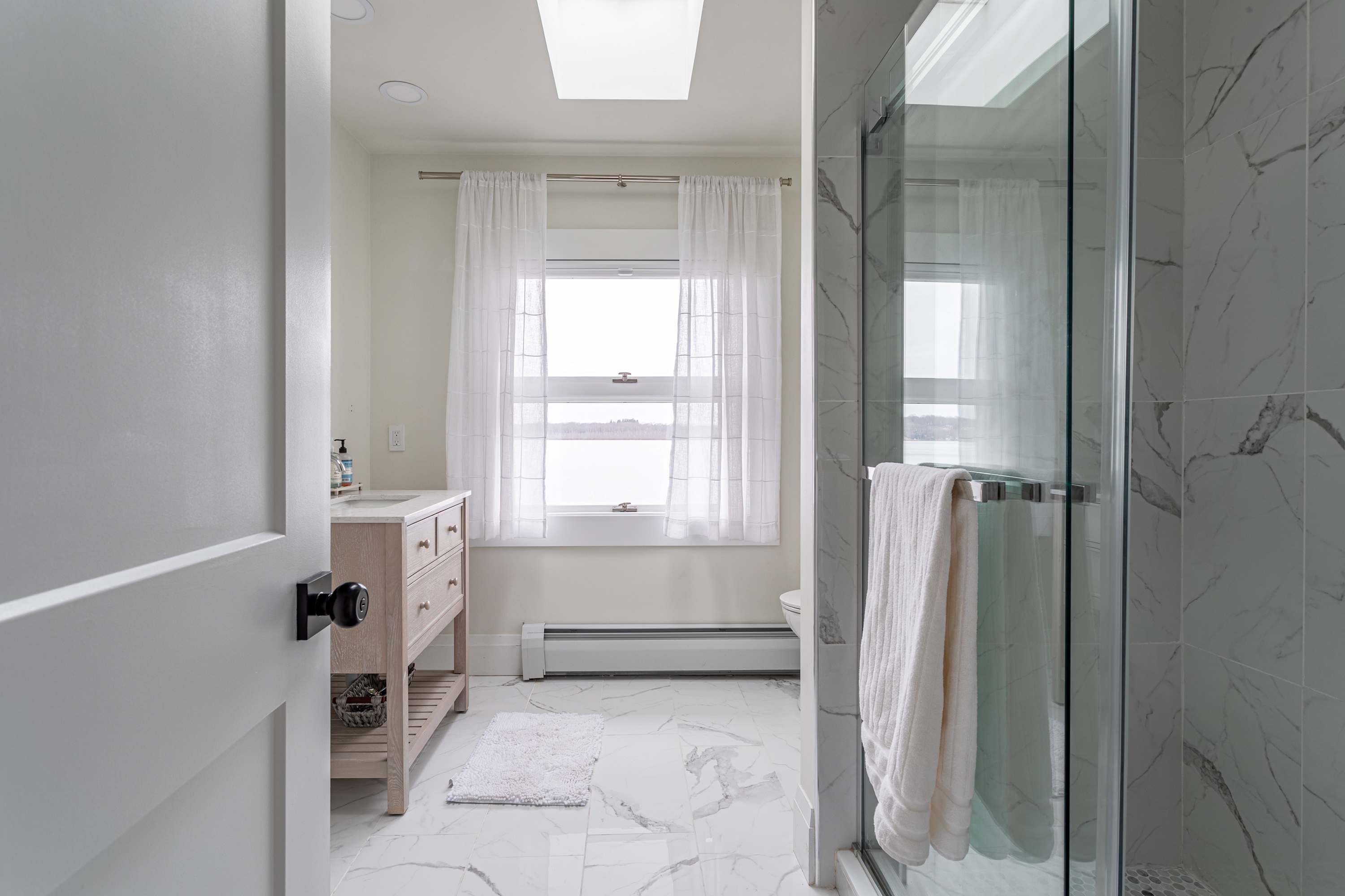 A bright bathroom with white marble-patterned floor tiles, a light wood vanity, and a window with white curtains offering a view of the lake. A skylight is visible above.