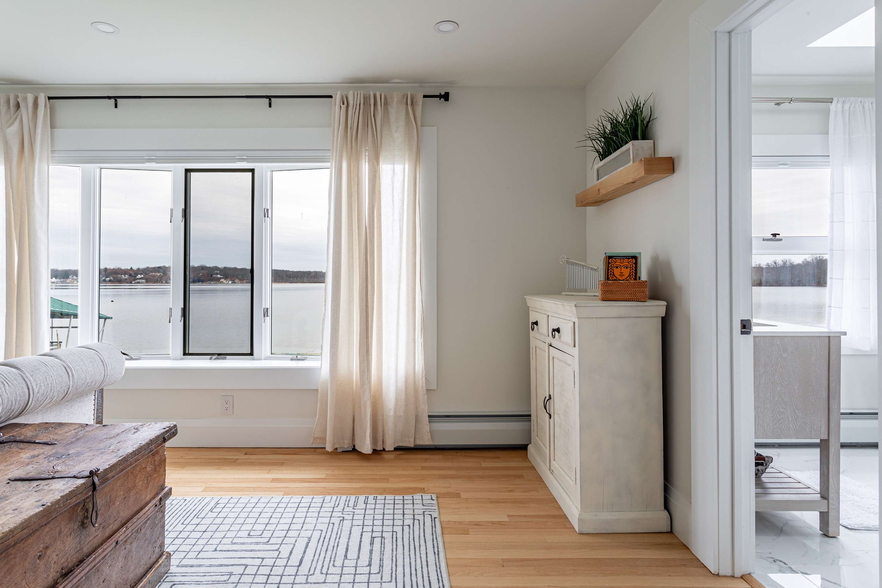 A bright bedroom with light hardwood floors and large windows offering a panoramic view of a lake. A white distressed cabinet with decor sits against the wall, and a patterned rug is on the floor.