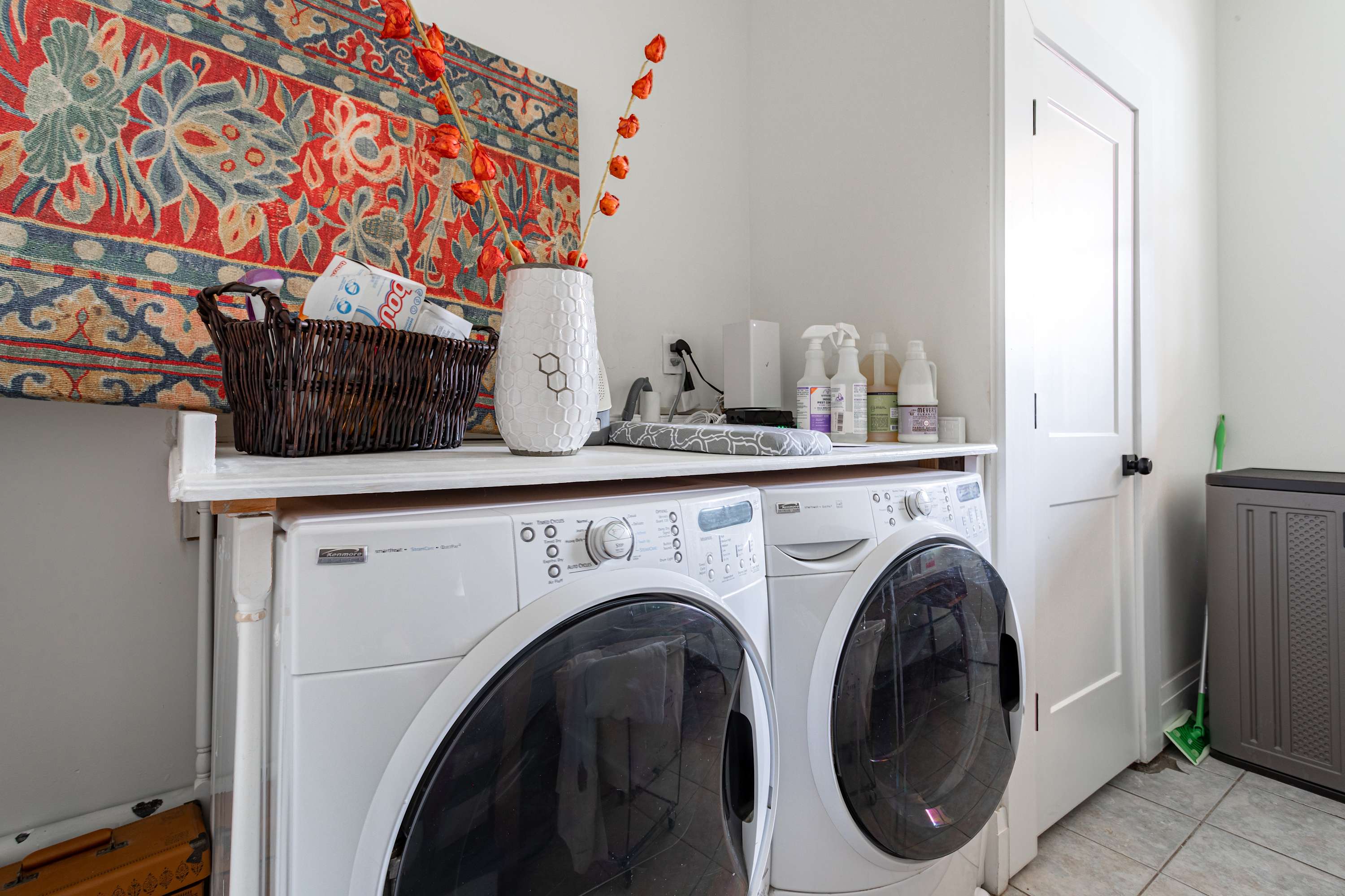A functional laundry area with a white front-loading washer and dryer set, topped by a shelf holding cleaning supplies and a decorative basket. A colorful tapestry hangs on the wall.