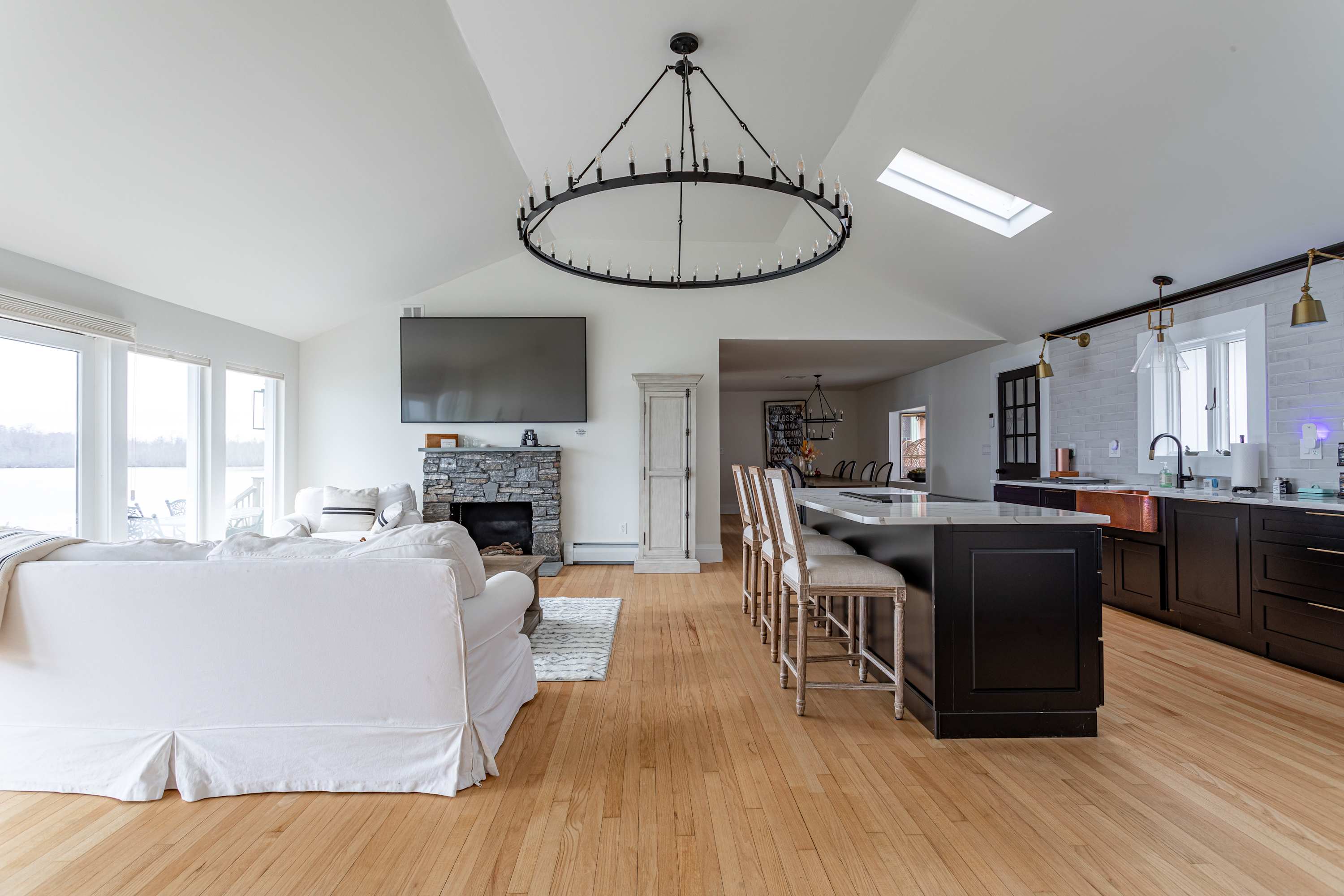 Wide view of the open-concept living area and kitchen, showcasing a white sofa facing a stone fireplace and wall-mounted TV, with a large kitchen island and dark cabinetry visible under a vaulted ceiling with a circular chandelier. Large windows offer lake views.