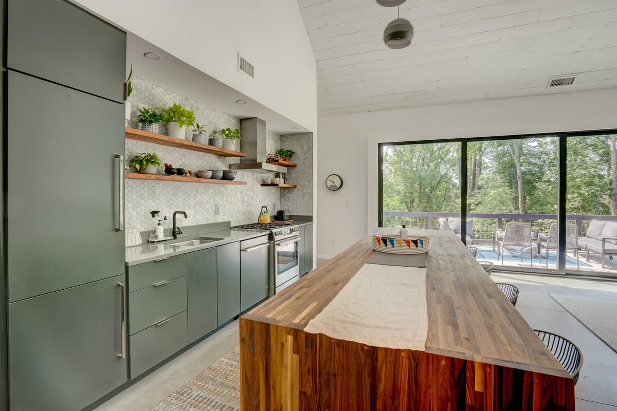 A modern kitchen boasts dark grey cabinetry, a long butcher block island, and a white hexagonal tile backsplash, with large sliding glass doors opening to an outdoor deck and forest views.
