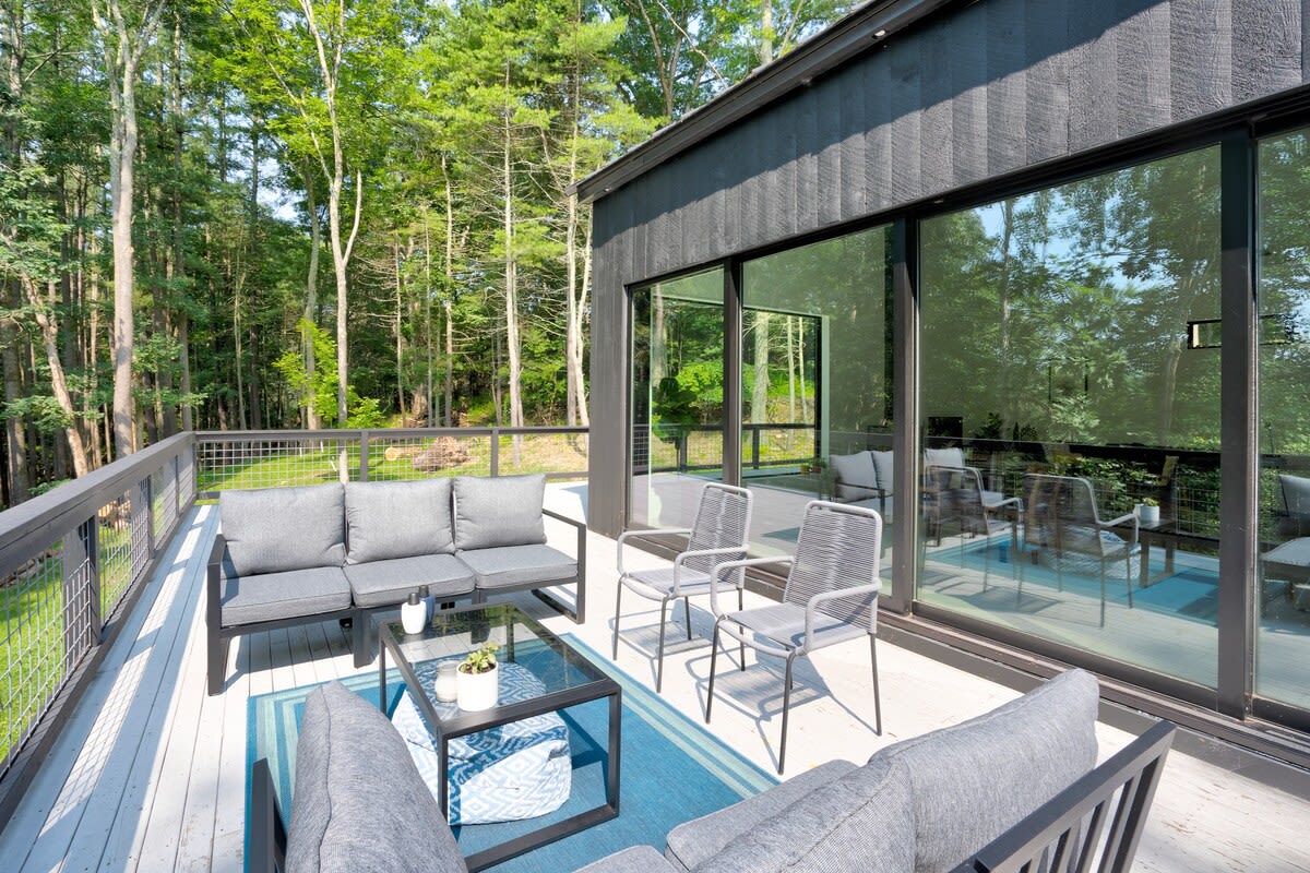 Expansive light-colored deck with modern gray outdoor seating, overlooking a lush forest, adjacent to the black modern house with large reflective glass windows and a metal railing.
