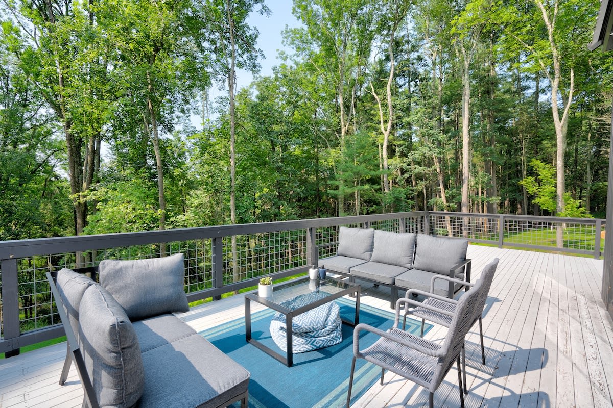 Spacious outdoor deck furnished with gray modular sofas and chairs, a glass-top coffee table, and a blue outdoor rug, all set against a backdrop of dense green trees and a metal railing.