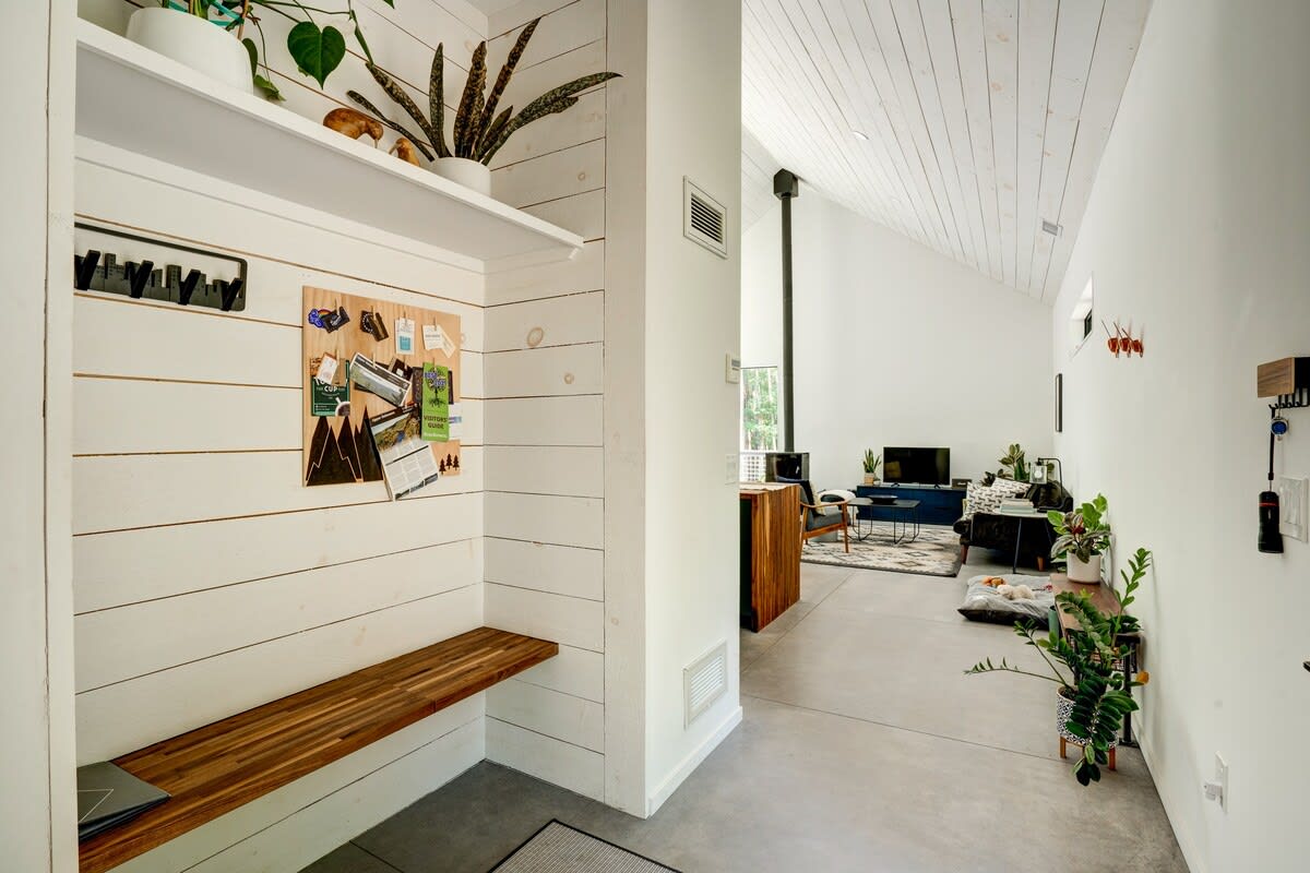 A modern foyer features white shiplap walls, a wooden bench, and a shelf with plants, leading into an open-concept living area with concrete floors and a vaulted ceiling.