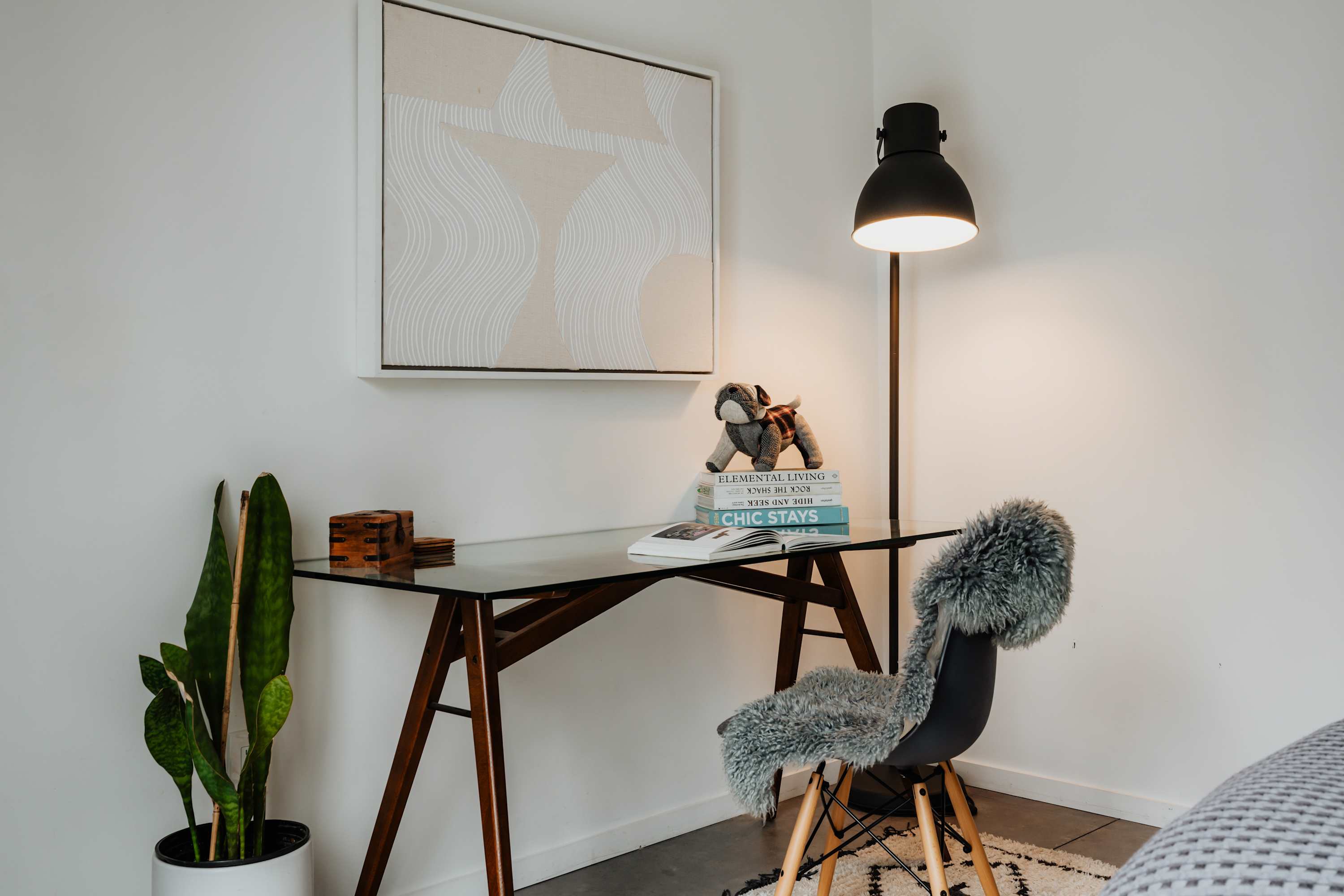 Cozy office nook featuring a glass-top desk with wooden legs, a black chair with a furry throw, a tall black floor lamp, and abstract artwork on a white wall, with a snake plant nearby.