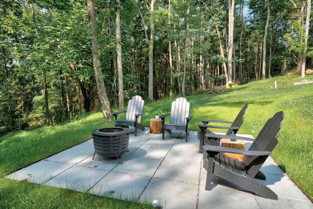 Ground-level fire pit area with a black metal fire pit and four black Adirondack chairs arranged on a square stone patio, surrounded by a lush green grassy slope and tall trees.
