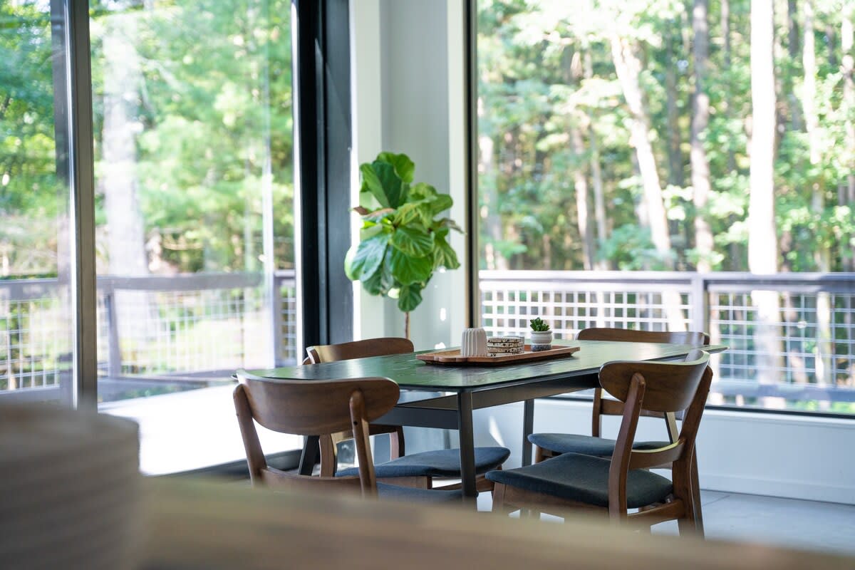 A bright dining area features a dark rectangular table with wooden chairs, positioned by expansive floor-to-ceiling windows that offer a lush view of the surrounding forest.
