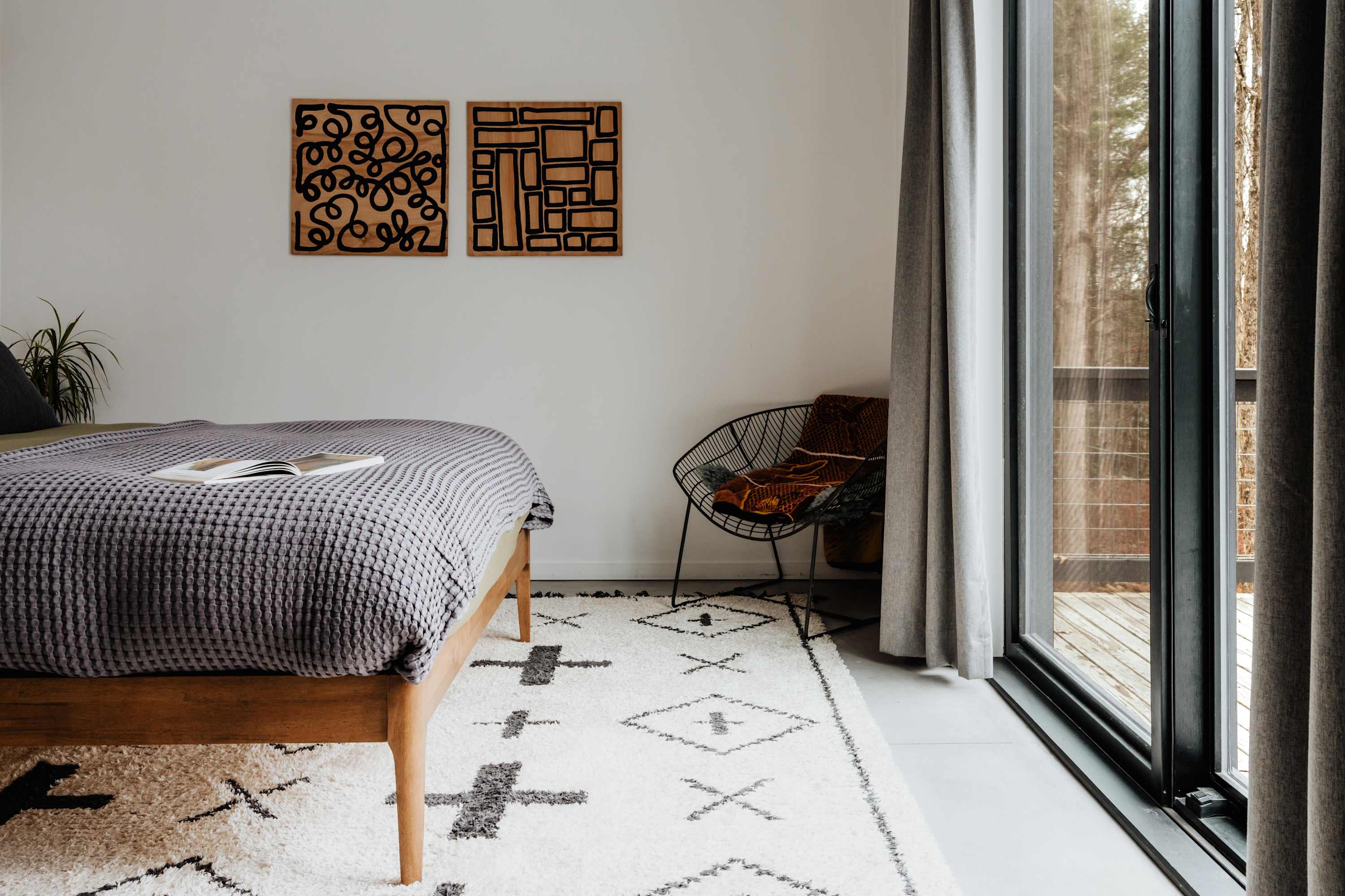 A modern bedroom features a wooden bed frame with a gray textured blanket and two abstract wooden art pieces on a white wall. A black wire chair with a patterned throw blanket sits beside a large glass door leading to an outdoor area, complemented by a white shag rug with black geometric patterns.