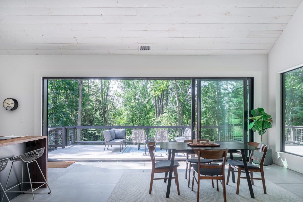 Bright dining area with a dark rectangular dining table and wooden chairs, featuring expansive sliding glass doors that open to a deck with lush green forest views, under a white vaulted ceiling.