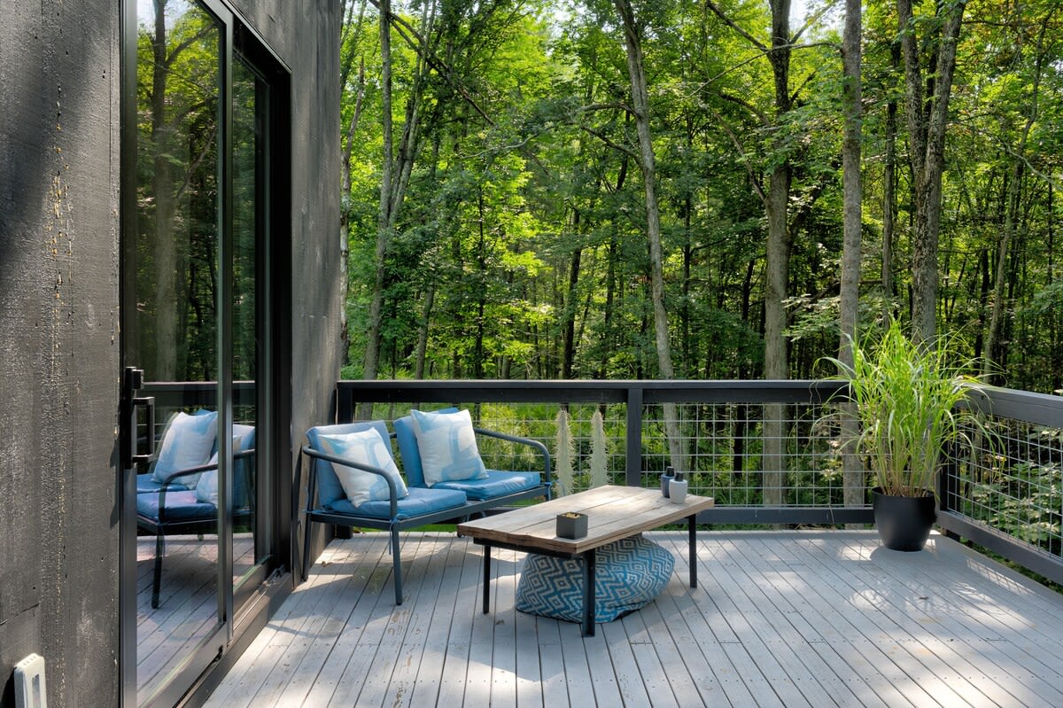 A spacious outdoor deck with light gray wooden planks features a blue metal bench with light blue cushions and a rustic wooden coffee table. A large potted plant adds greenery, and the deck is surrounded by lush trees, accessible via a large glass sliding door.