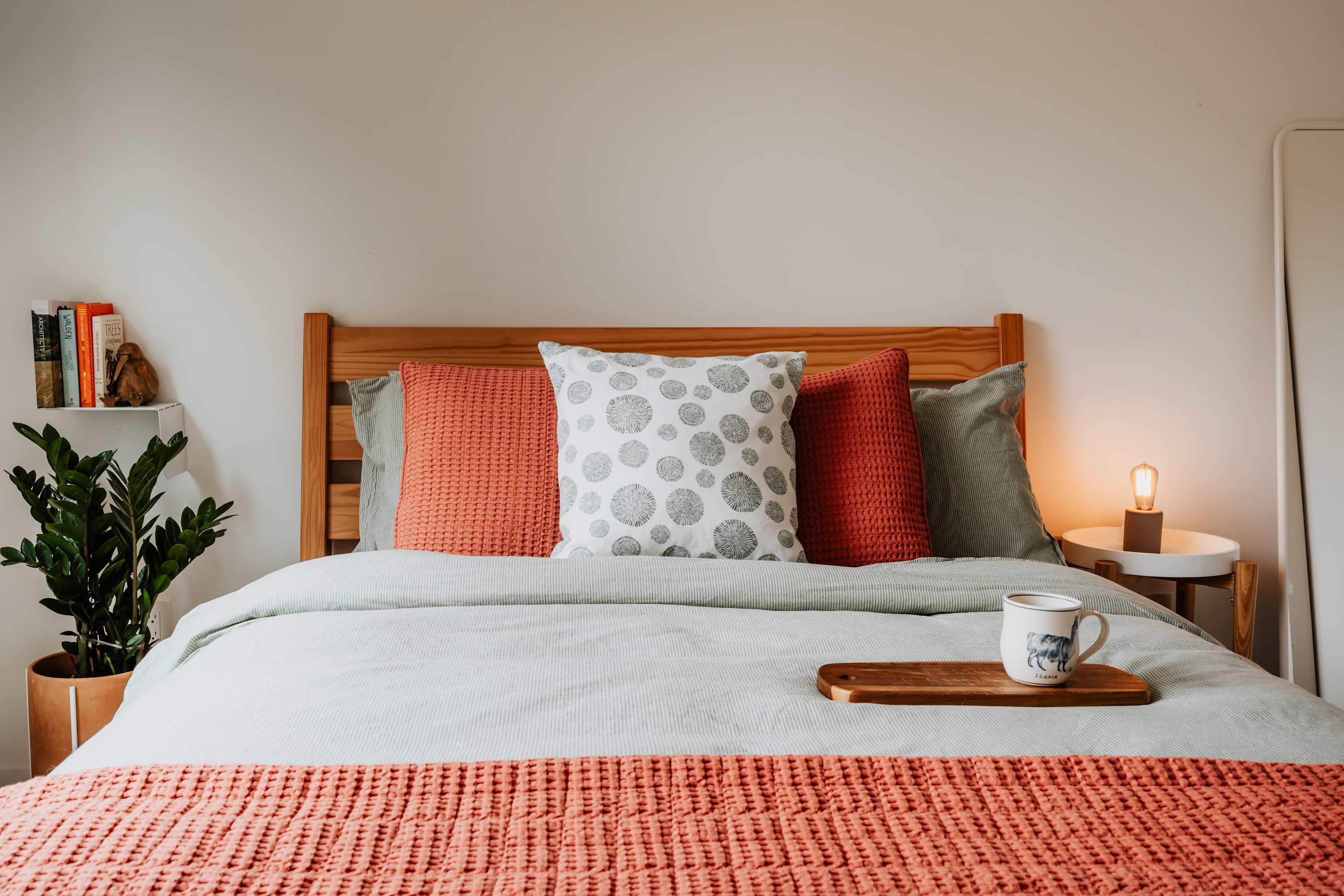 A cozy bedroom features a light wooden bed frame dressed with light green and coral bedding, accented by decorative pillows, a small side table with a lamp, and a potted plant on a shelf.